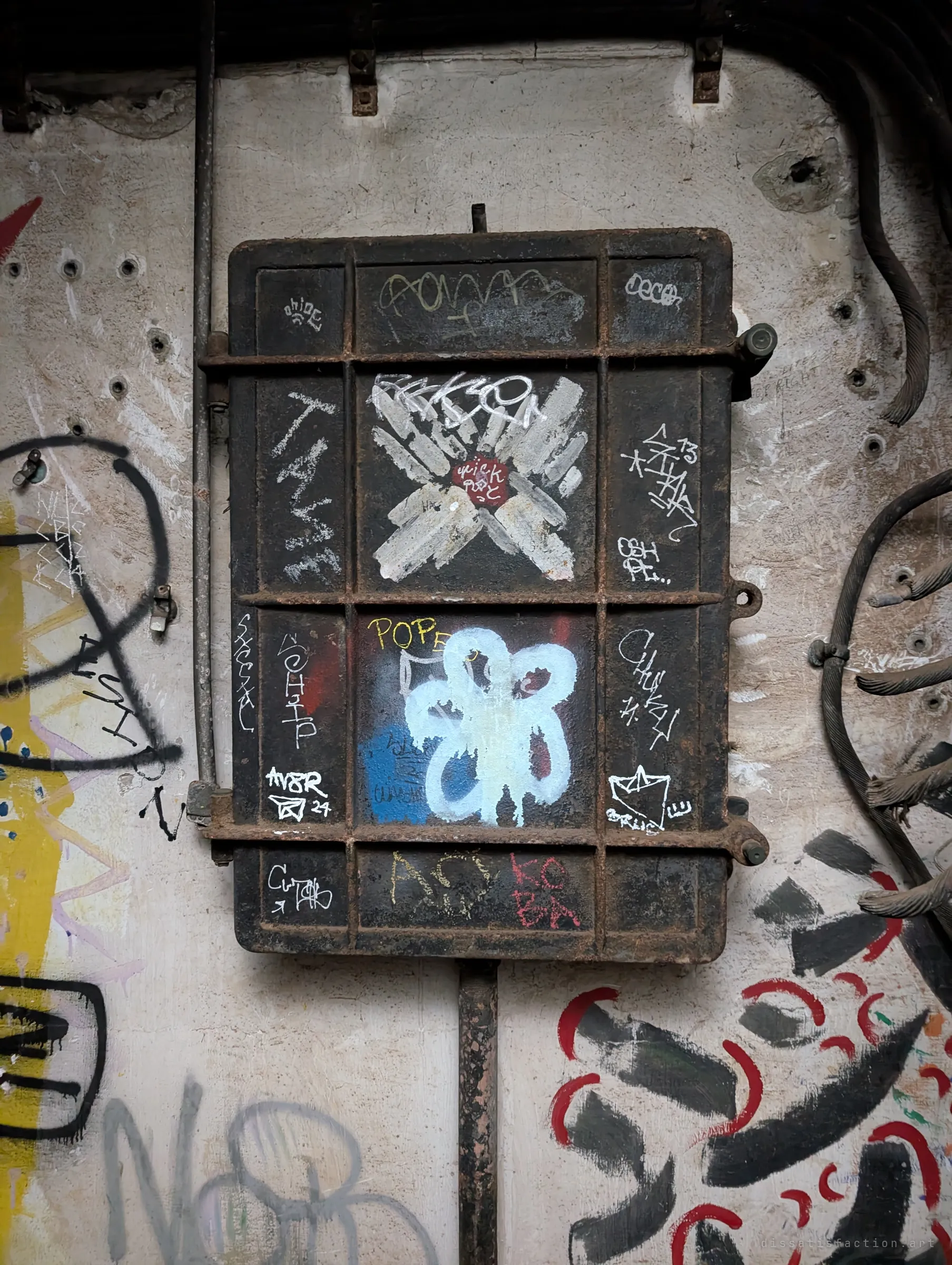A heavy rusted iron panel box bolted to a concrete wall, its door covered in small tags and a white painted flower. Thick severed cables curl out of the wall beside it.