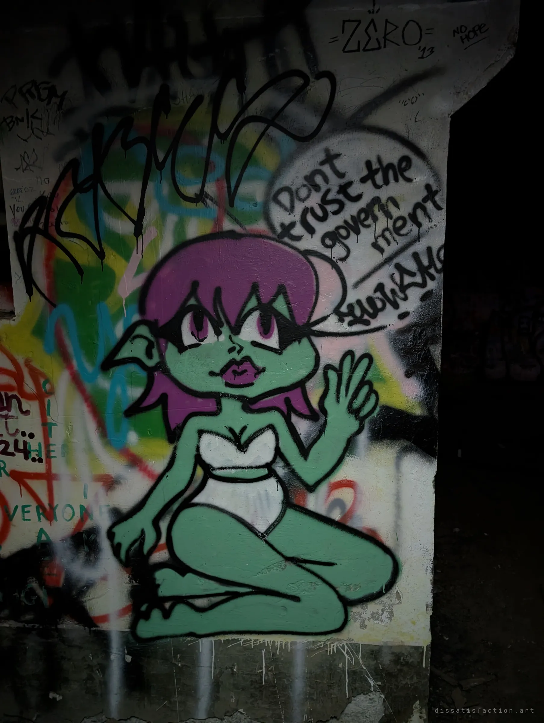 A painted green goblin girl with purple hair waves beside a speech bubble warning not to trust the government, surrounded by older tags in the dark. One of the more characterful pieces in the tunnels.