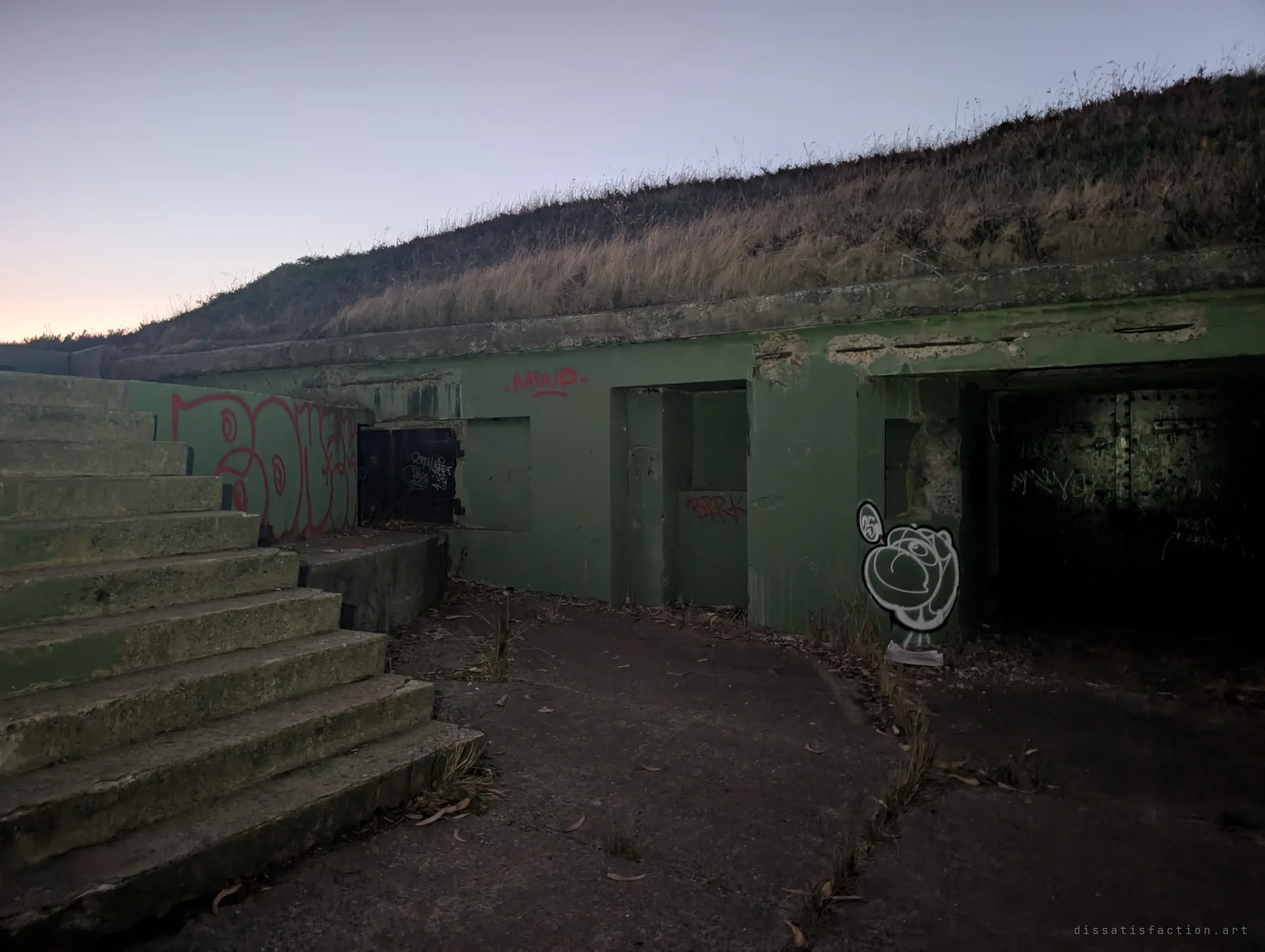 A green concrete bunker set into a grassy earth mound, with worn steps on the left and dark doorways marked with graffiti. The earth cover was built to hide and shield the structure.