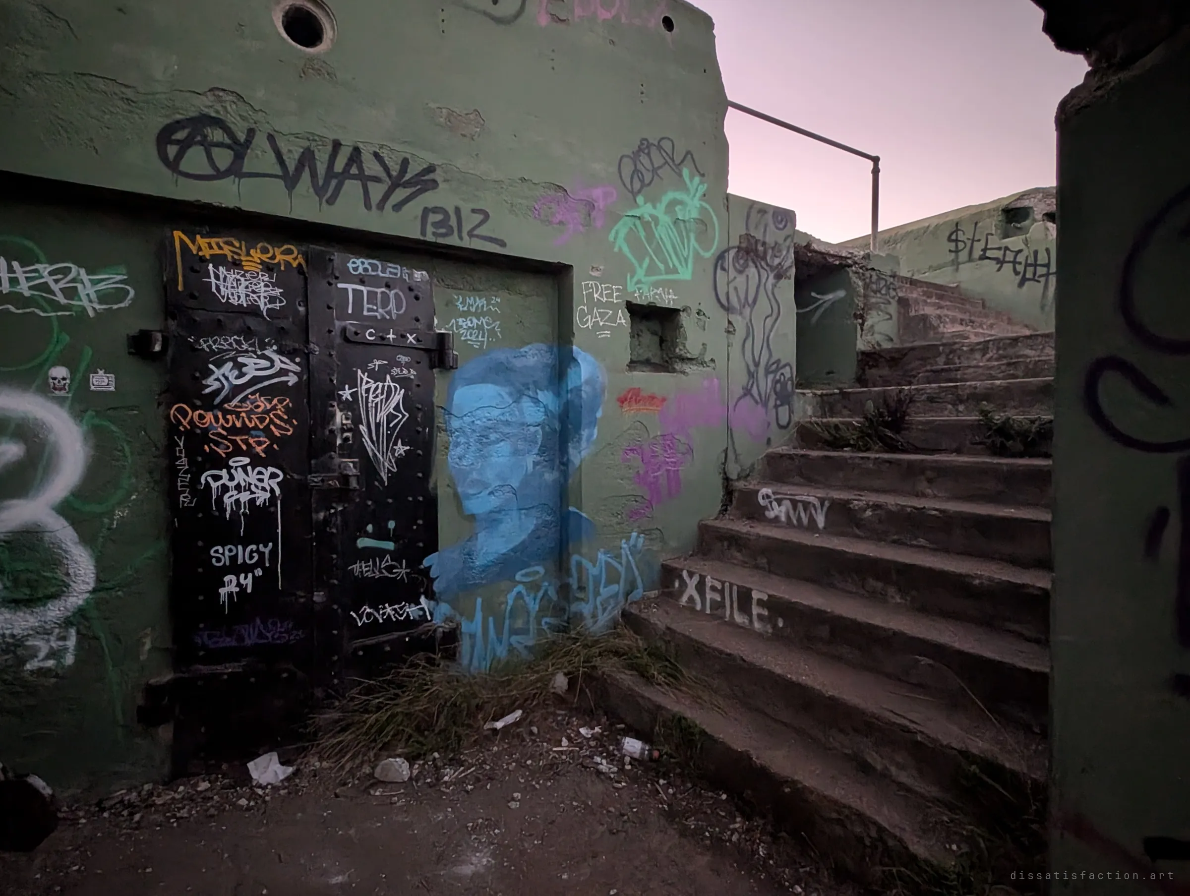 A riveted steel double door covered in tags beside a pale blue painted portrait, with a weathered concrete stair climbing to the right under a dusk sky. Layers of graffiti cover the old military concrete.
