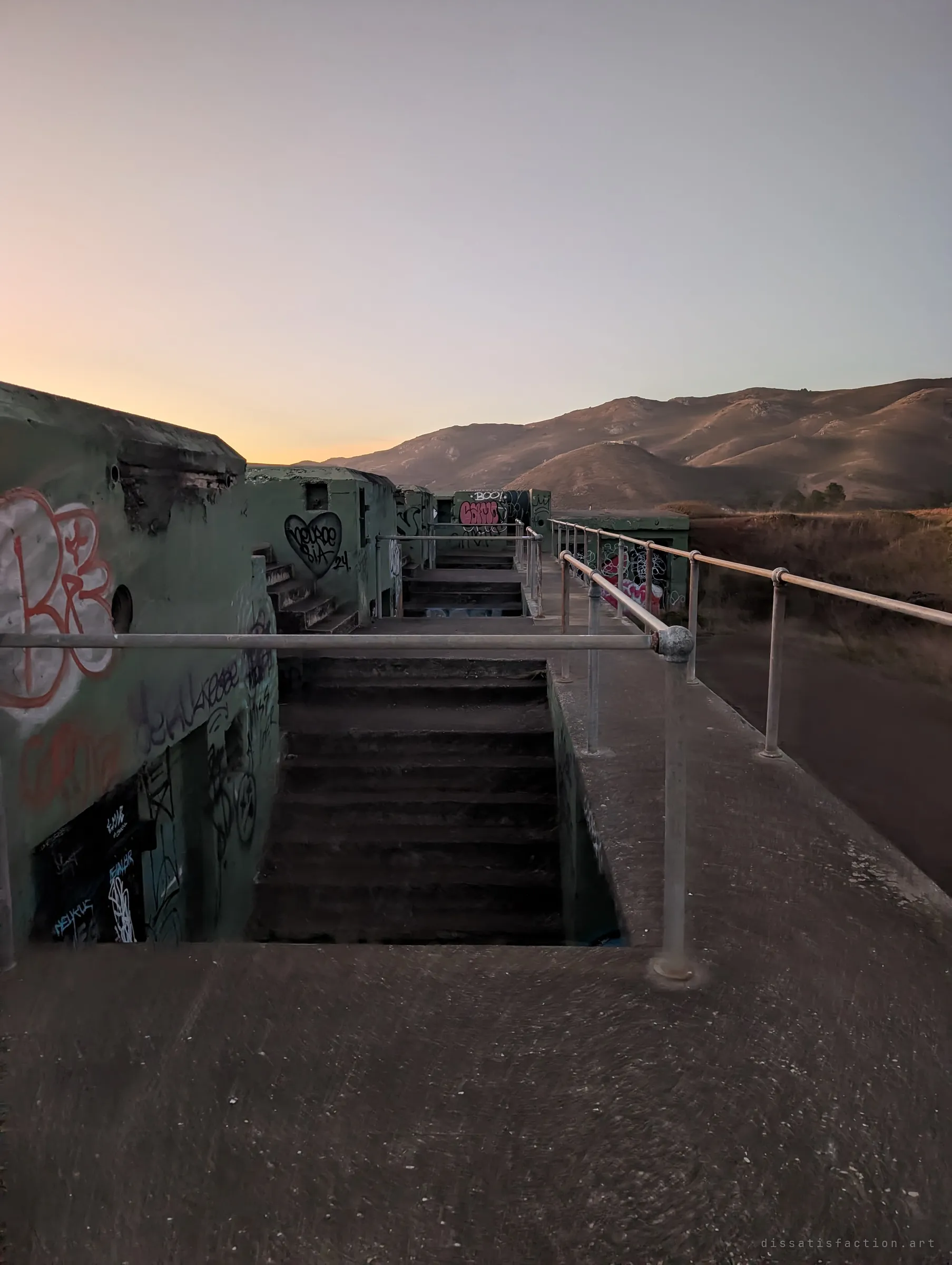 Stairs drop into a graffiti-covered concrete gun emplacement, railings running toward bare golden hills under a dusk sky. The fort's layered stairwells and walkways show how the battery was stepped into the slope.