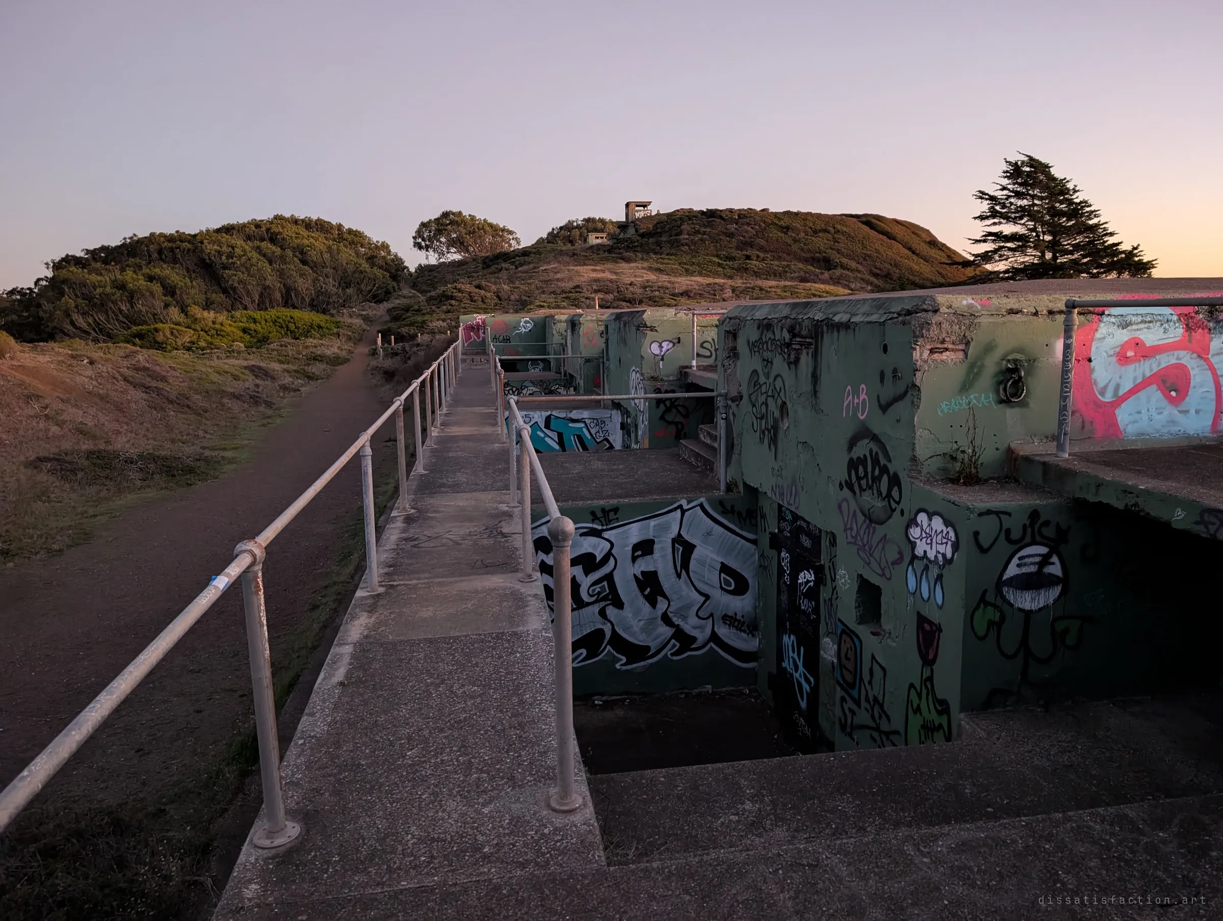 A railed concrete walkway runs along a row of graffiti-covered emplacements, with a small observation post on the scrubby hill beyond. Dusk light catches the tops of the bunkers.