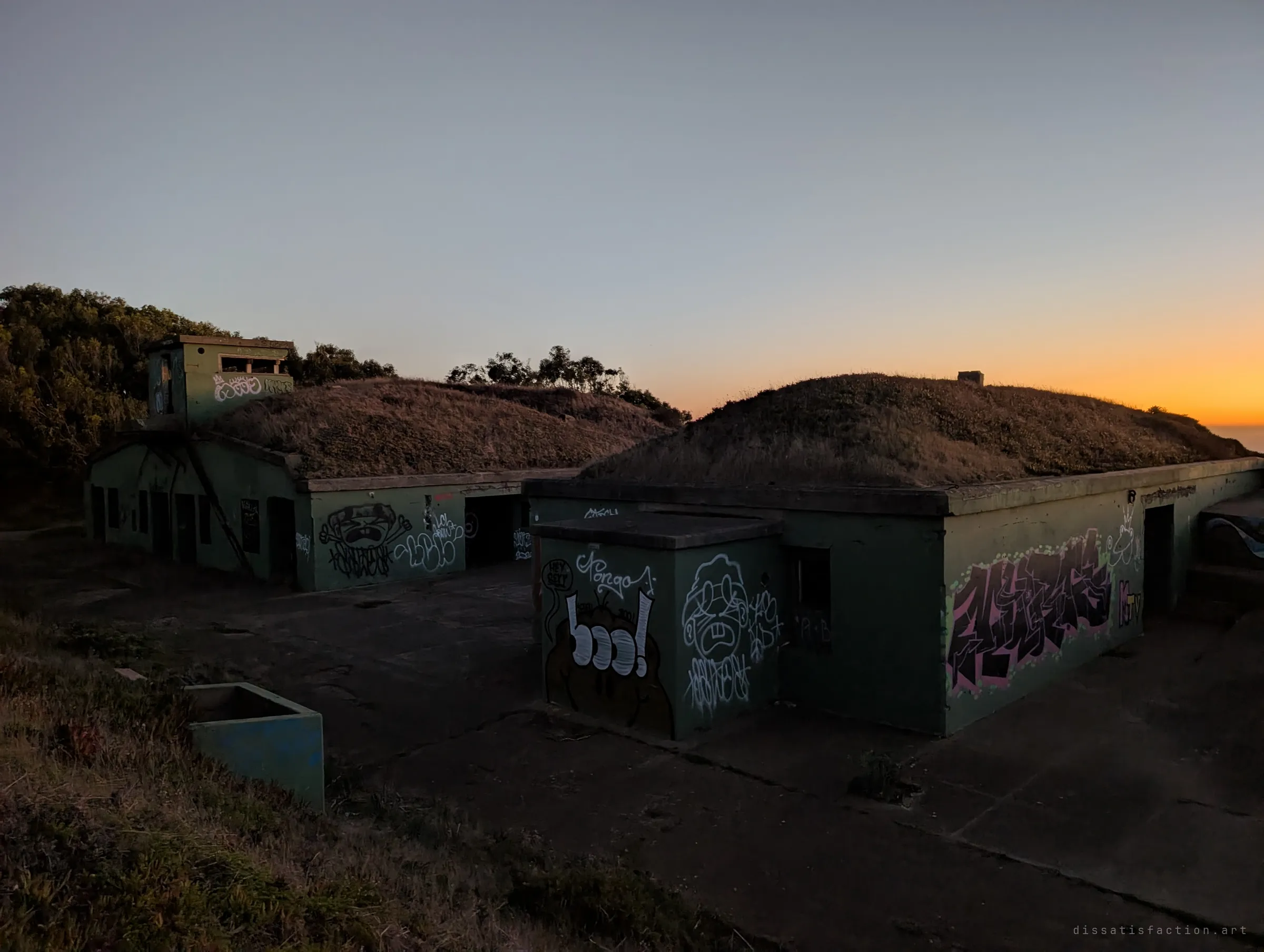 Two grass-roofed concrete bunkers covered in graffiti sit in shadow as the sun sets over the sea behind them. A small lookout tower rises from the left-hand mound.
