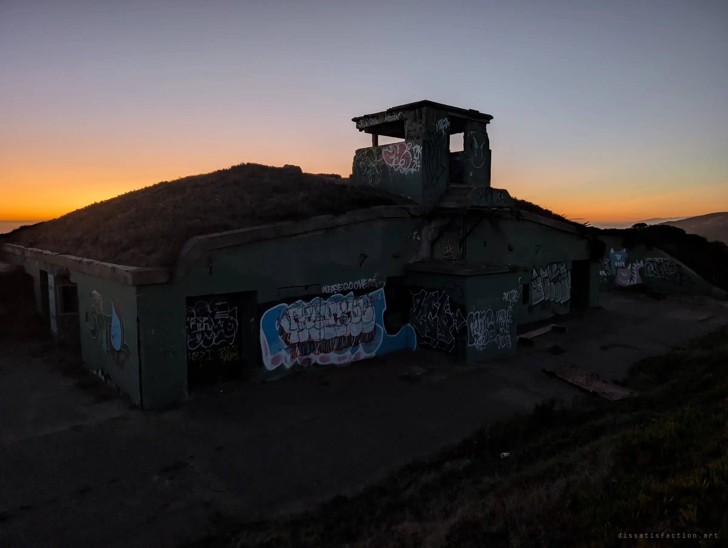 A graffiti-covered bunker with an open lookout on its roof stands silhouetted against an orange sunset over the ocean. The concrete apron in front is already in darkness.