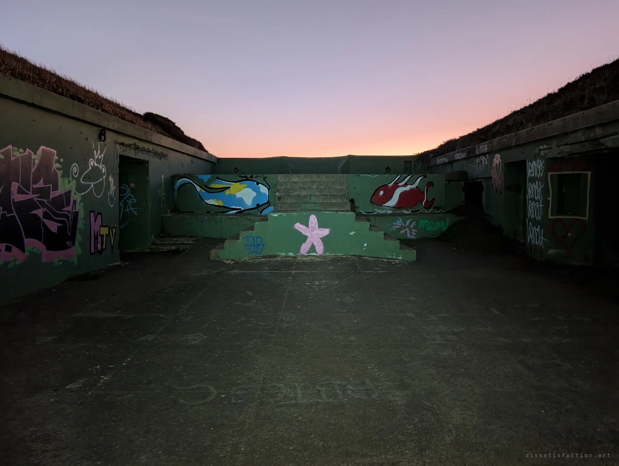 An open gun pit framed by earth-covered walls, with painted fish and a pink starfish on the central concrete steps. A pink-orange sky glows above the empty platform.