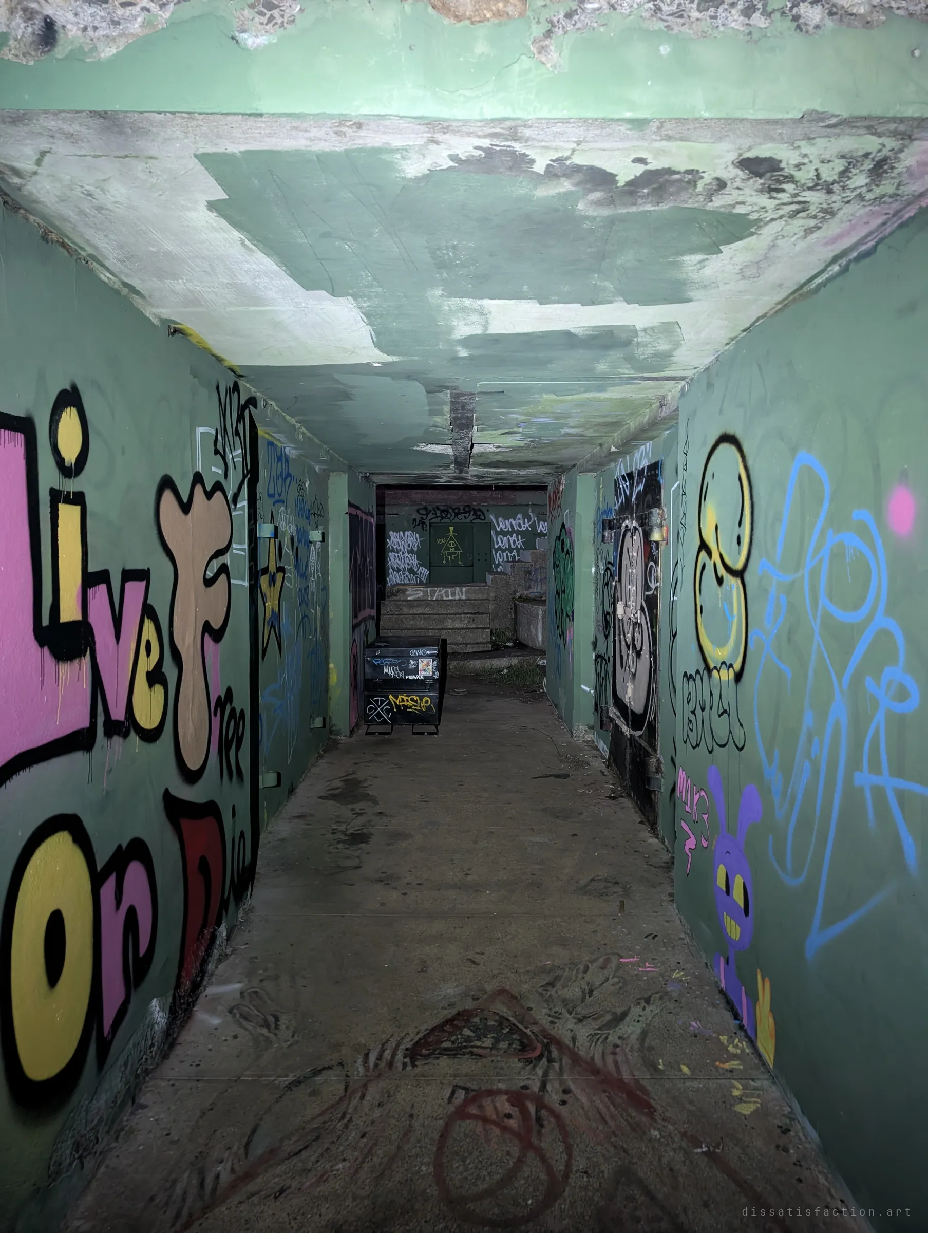 A narrow concrete corridor inside the battery, walls packed with bright graffiti and a peeling green ceiling. A metal bin and a short flight of stairs wait at the far end.
