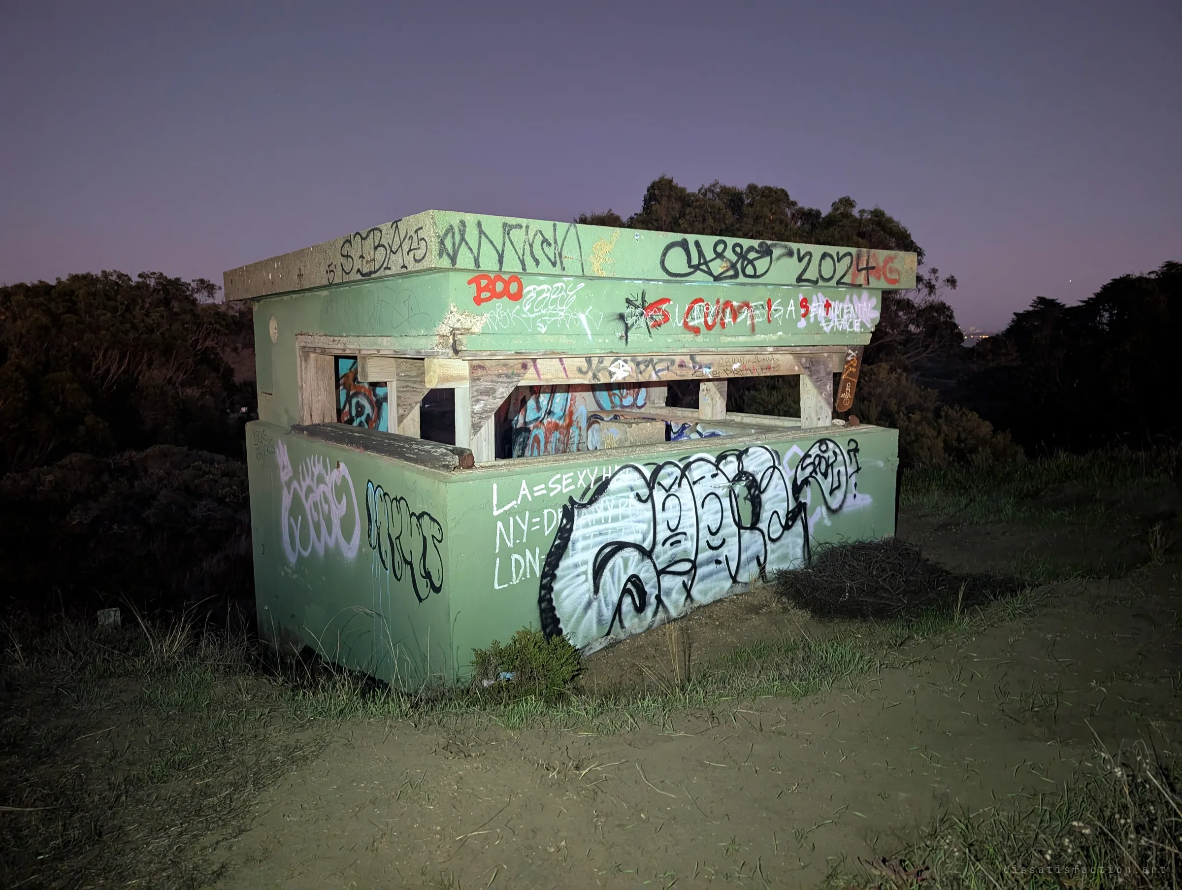 A small concrete observation post on a hilltop, lit by flashlight and layered with graffiti tags. Its timber-framed viewing slot faces out over dark scrub at dusk.