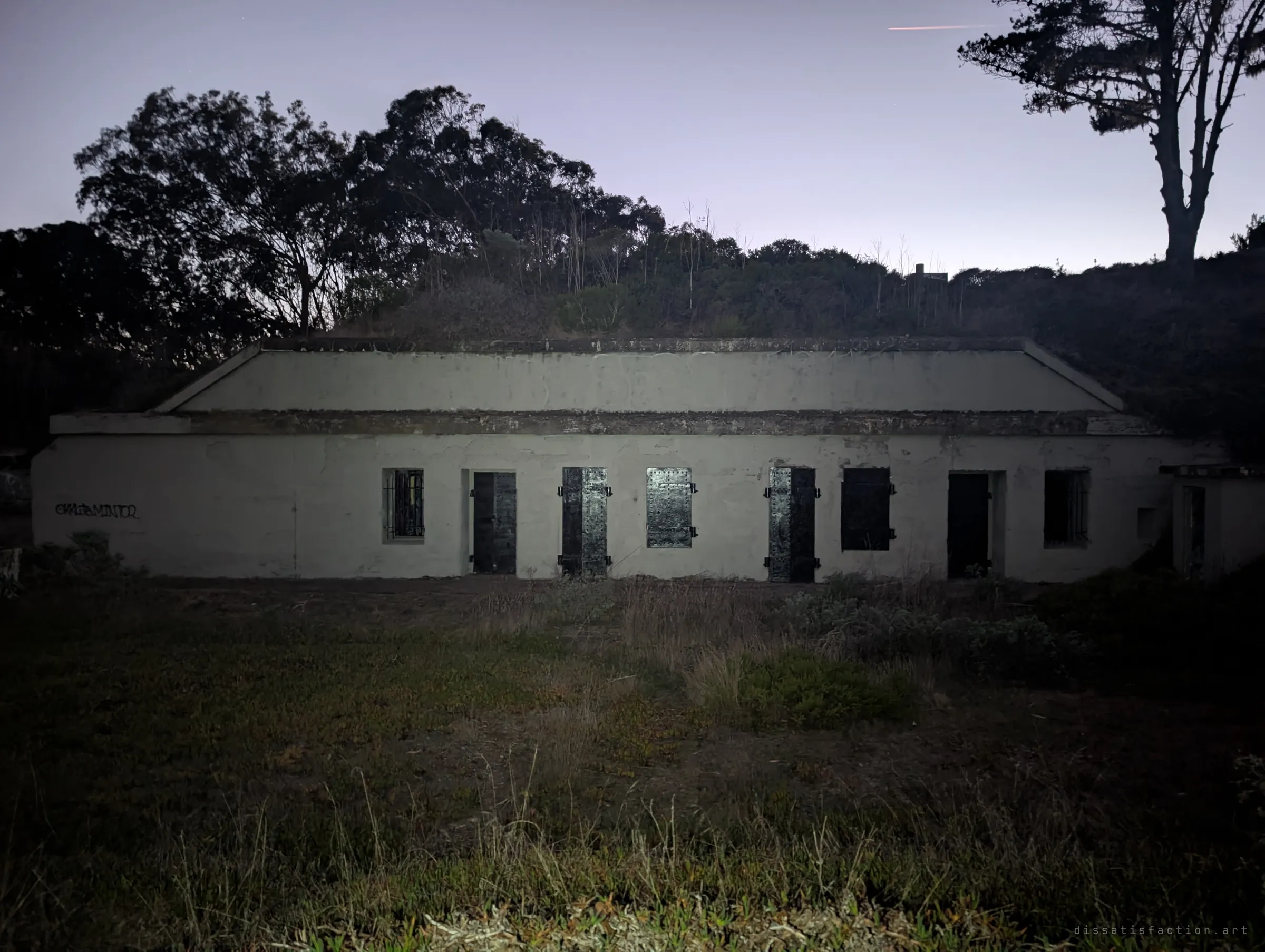 A long pale concrete building dug into the hillside, with a row of riveted steel doors and shutters. Trees silhouette the ridge above it at dusk.