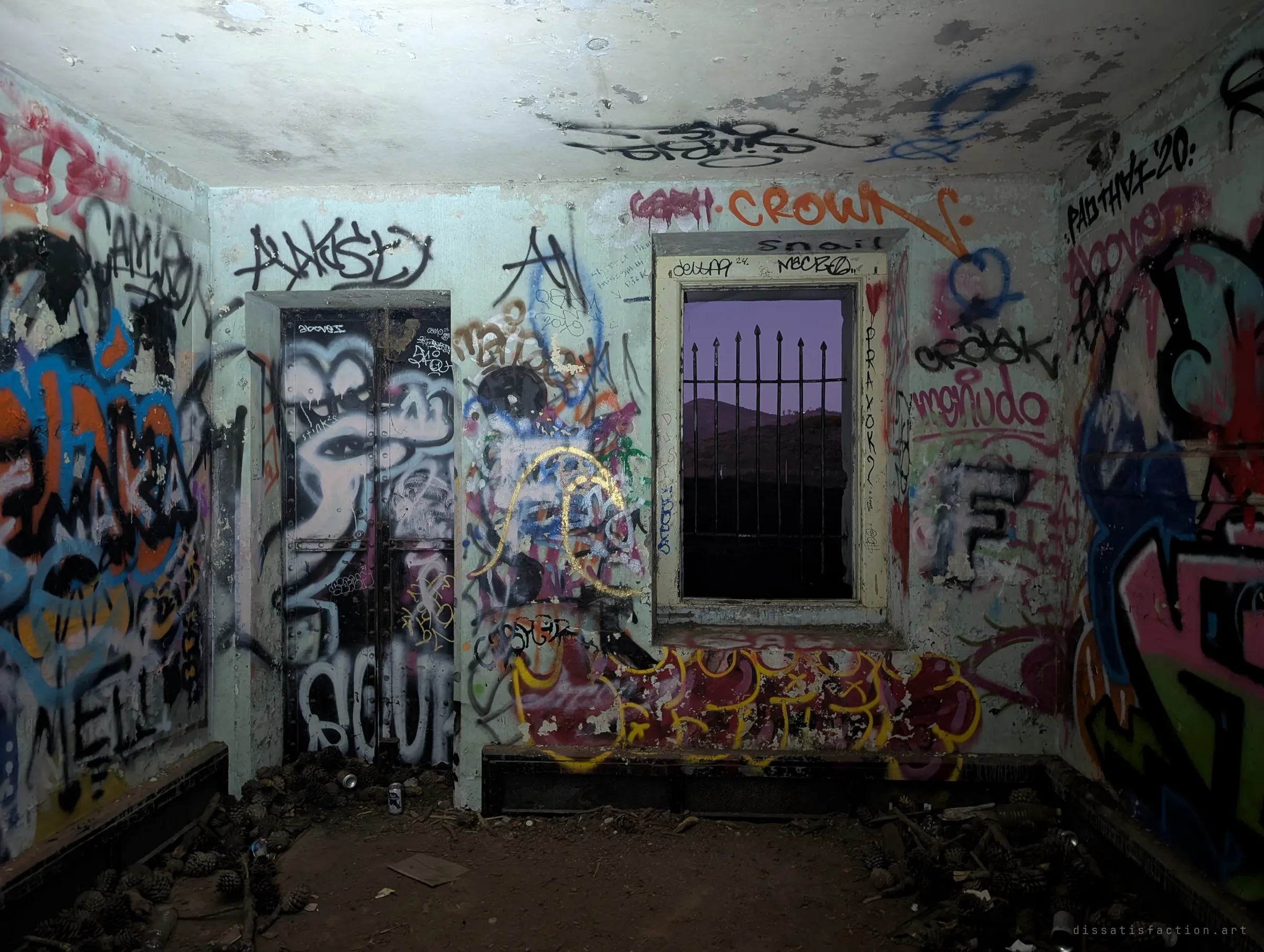 A small room inside the battery, every wall and the ceiling covered in graffiti, with pine cones and cans on the floor. A barred window frames a violet dusk sky over the hills.