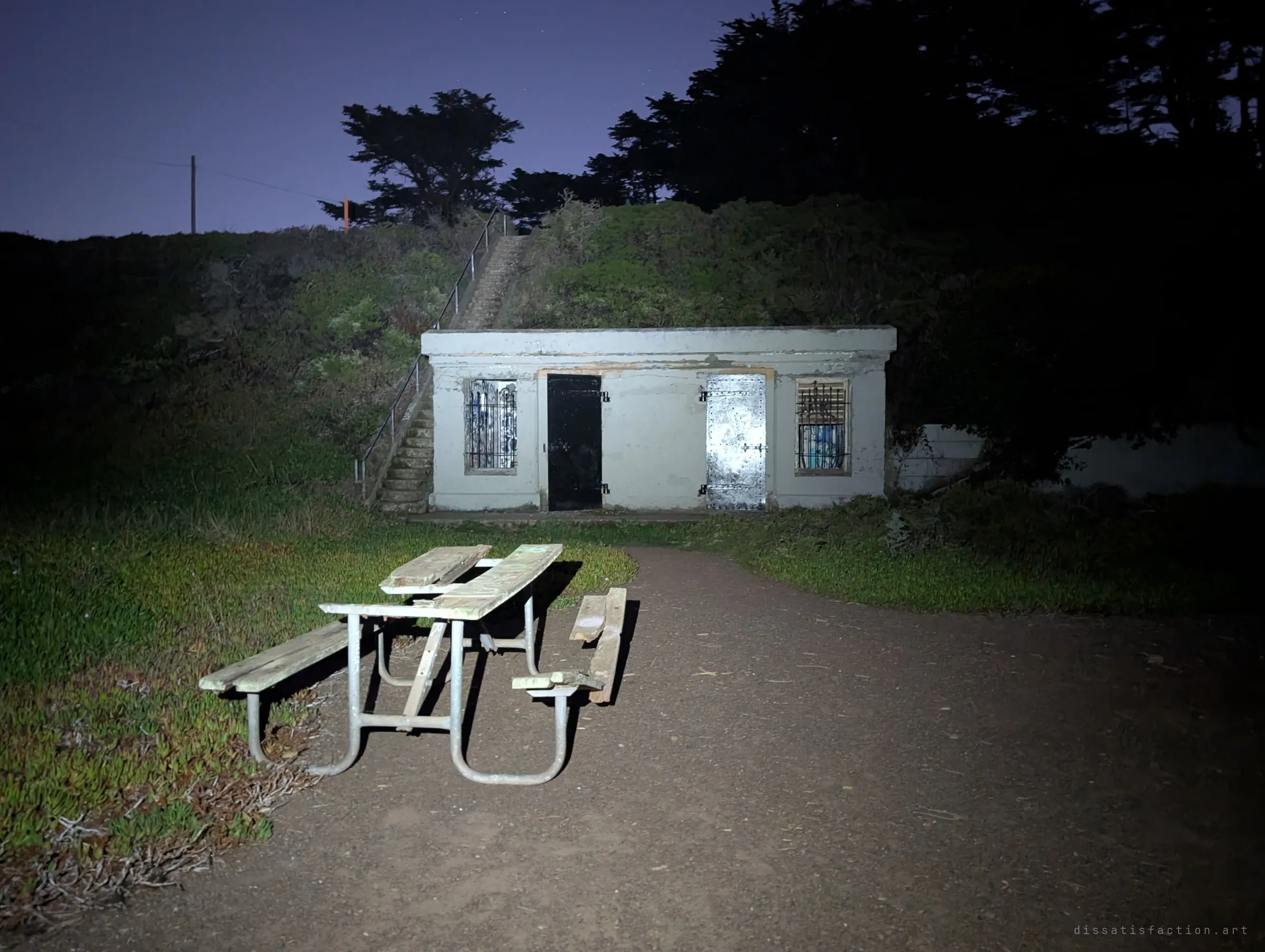 A broken picnic table sits in flashlight in front of a squat white bunker with barred windows and steel doors. Concrete stairs climb the grassy mound beside it into the night.