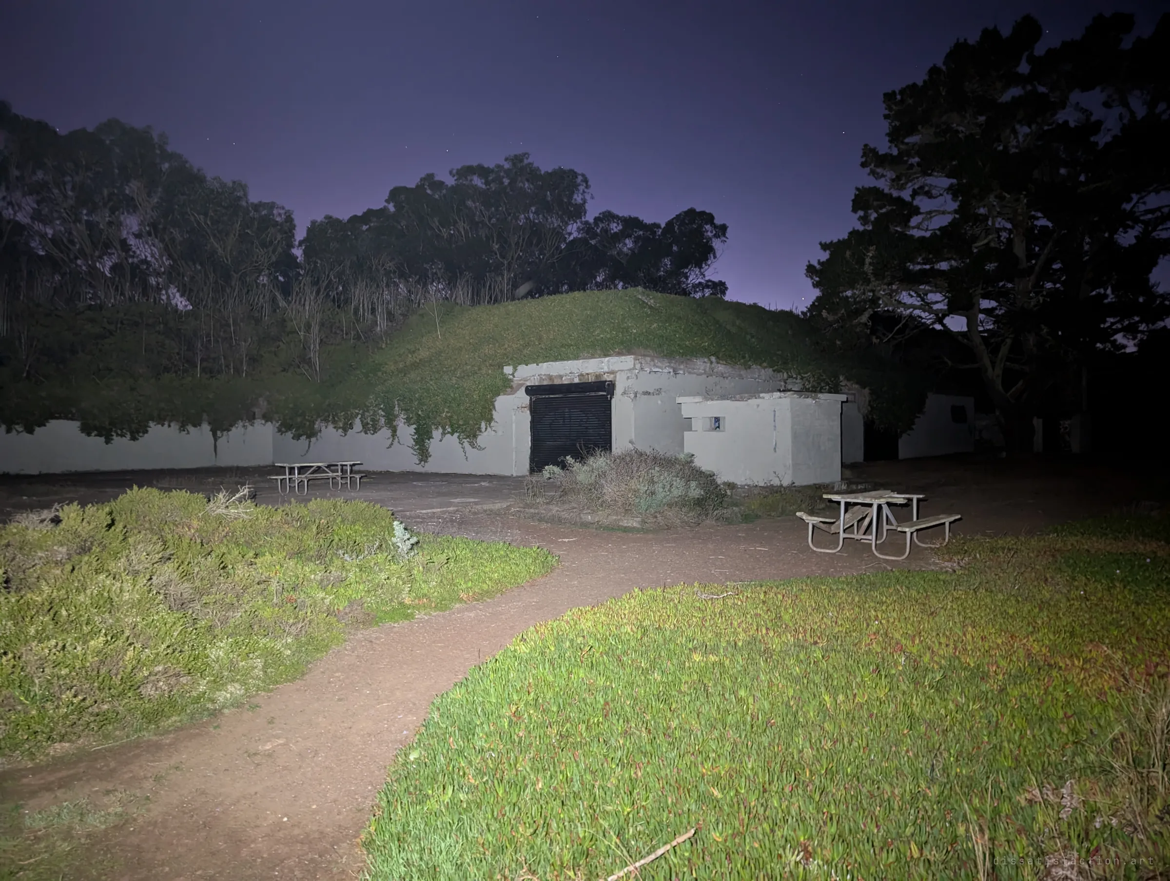 A dirt path curves across ice plant toward a grass-roofed bunker with a closed roller shutter and scattered picnic tables. Tall eucalyptus trees stand dark against the night sky.