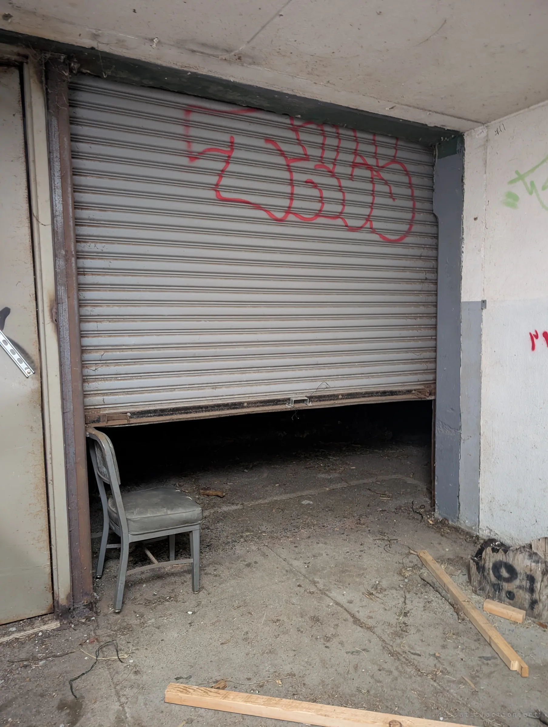 A grey roll-up shutter, tagged in red, stops a few feet above the floor, with an old metal office chair tucked under its edge and loose lumber on the concrete.