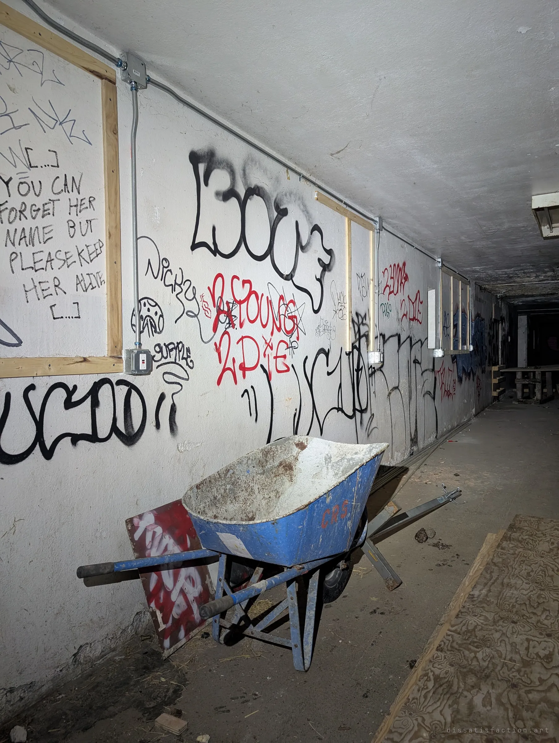 A long whitewashed corridor covered in tags and a handwritten plea about remembering someone, with a battered blue wheelbarrow parked against the wall and the passage fading into darkness.