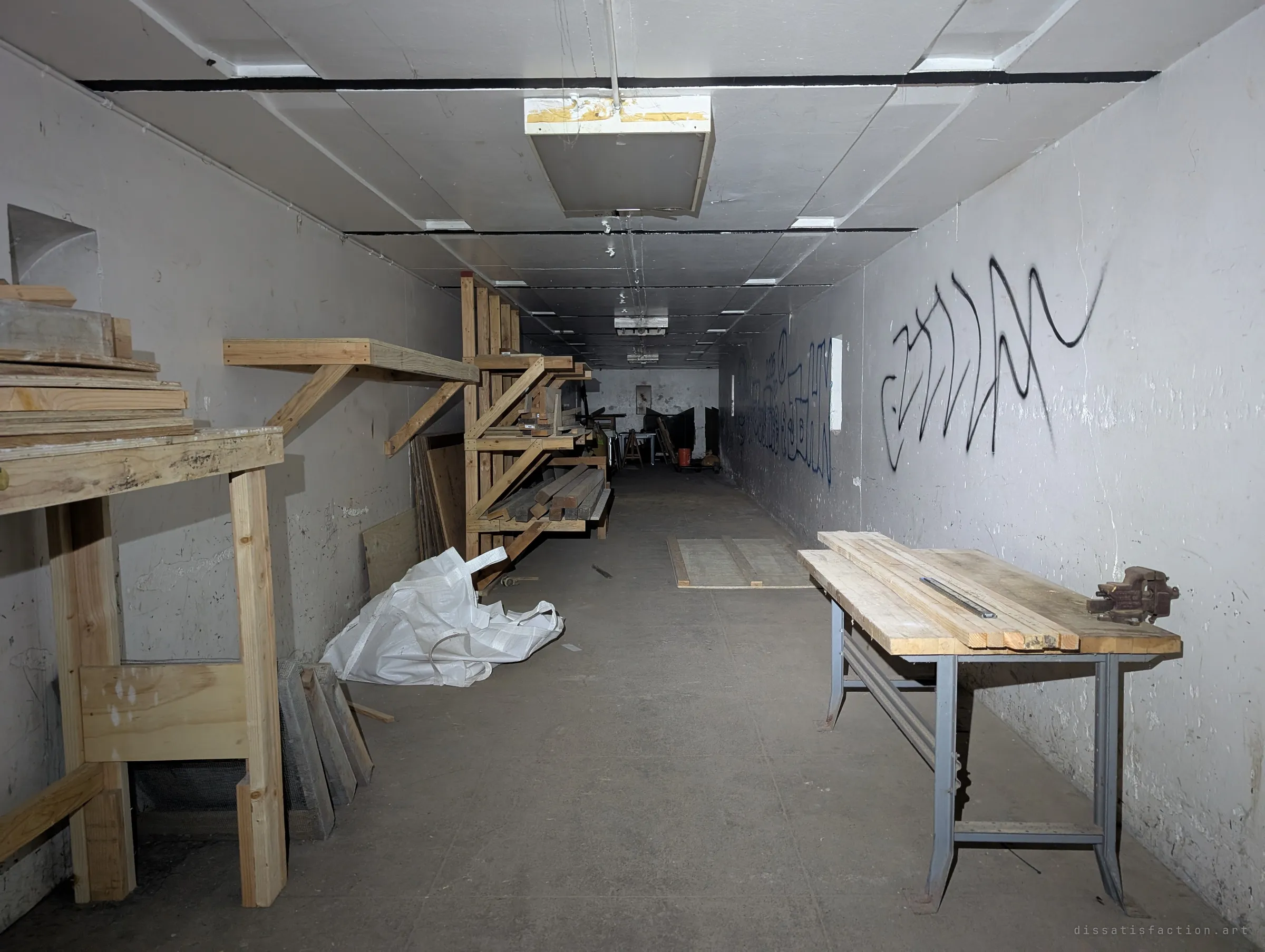 A long vaulted concrete storeroom turned workshop, with lumber racks and a workbench on the left and a bench with a vise on the right, running back toward more stored material.
