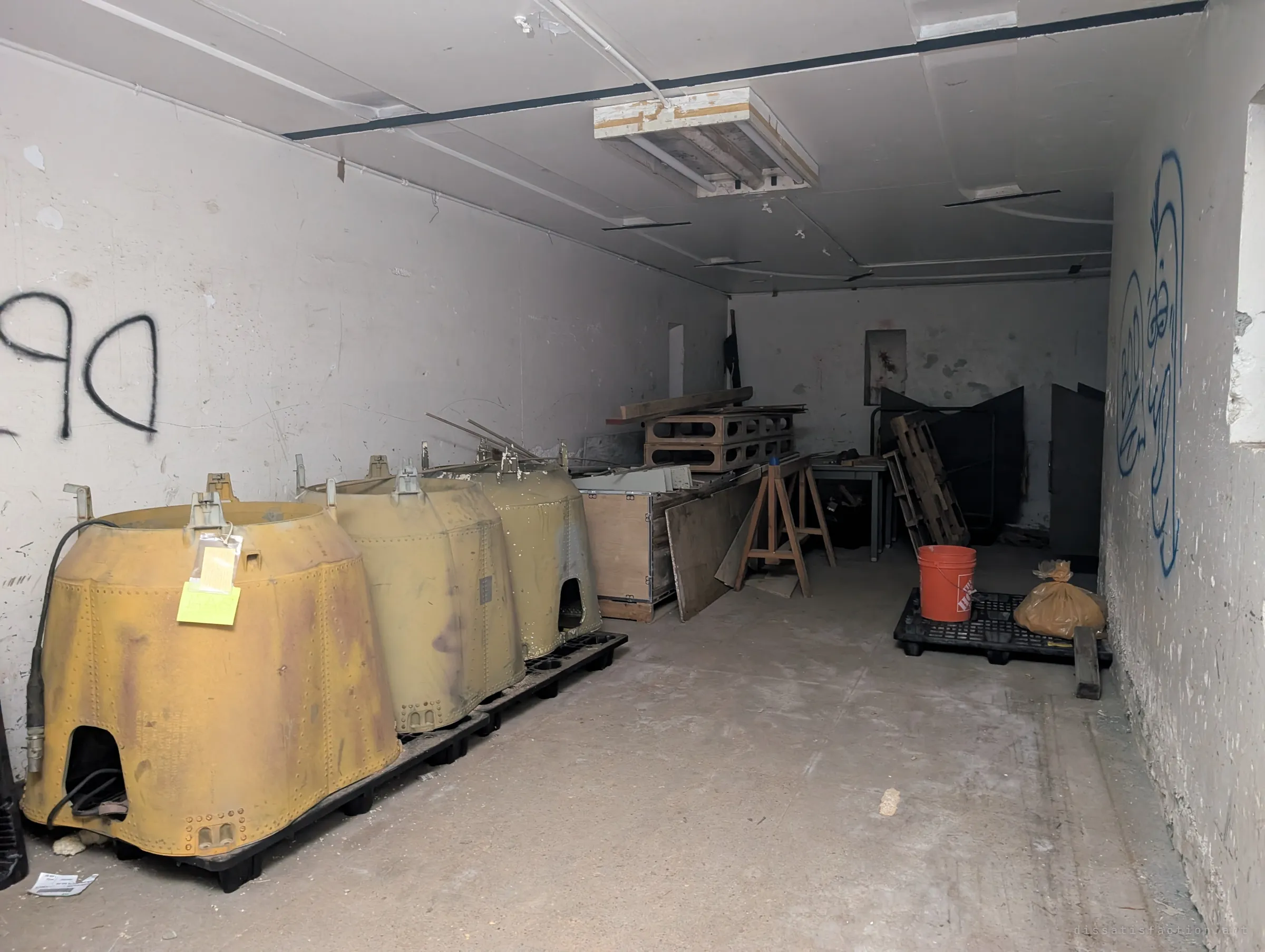 Three rusted yellow riveted metal housings sit on pallets in a white concrete room, tagged with inventory cards, beside crates, sawhorses and an orange bucket.