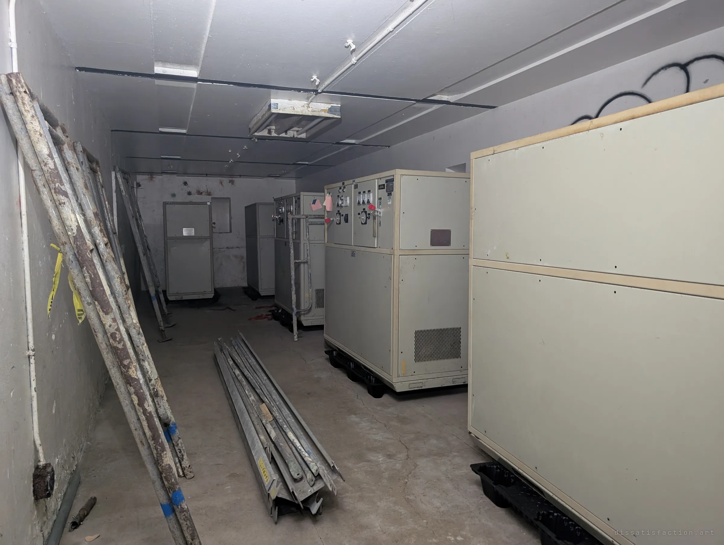 Rows of beige motor-generator cabinets with dials and switches stand in a narrow concrete room, with scaffolding poles leaning on the wall and piled on the floor.