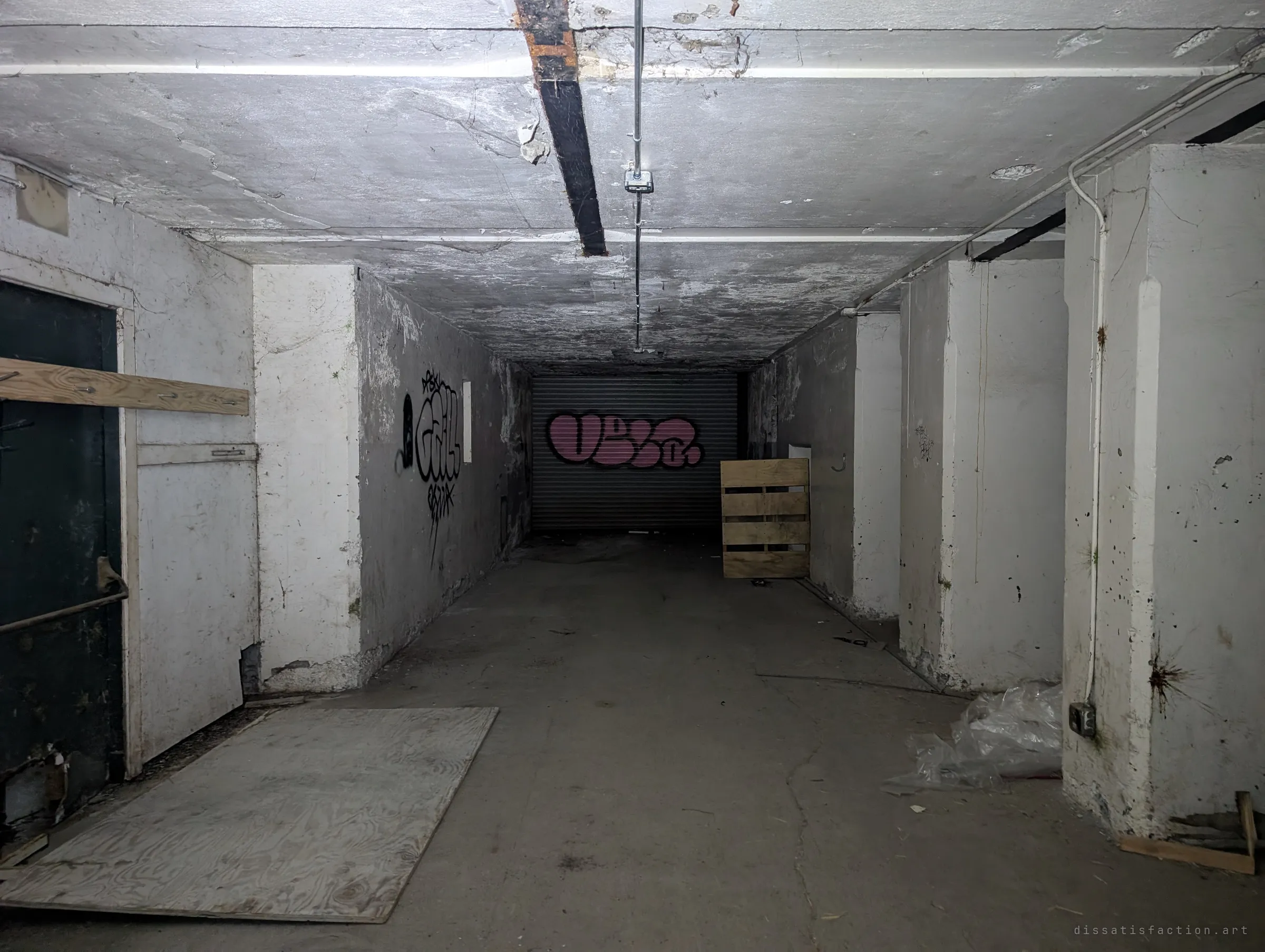 A heavy-walled concrete passage with thick pillars and a steel beam overhead leads to a pink-tagged roll-up door, a loose pallet propped against the side wall.