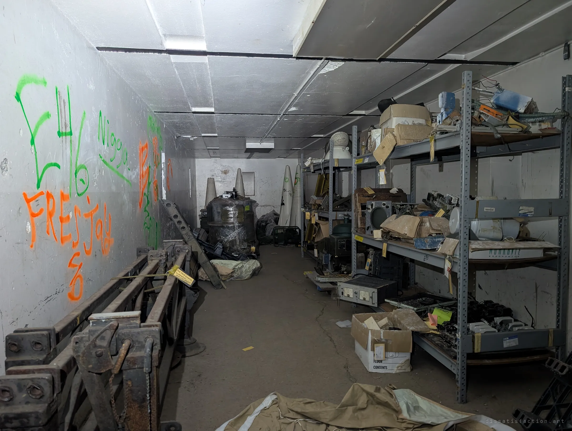 A storage gallery lined with steel shelves of tagged electronics and parts, rusted rail track sections on the left, and tall conical shapes wrapped in plastic at the far end.