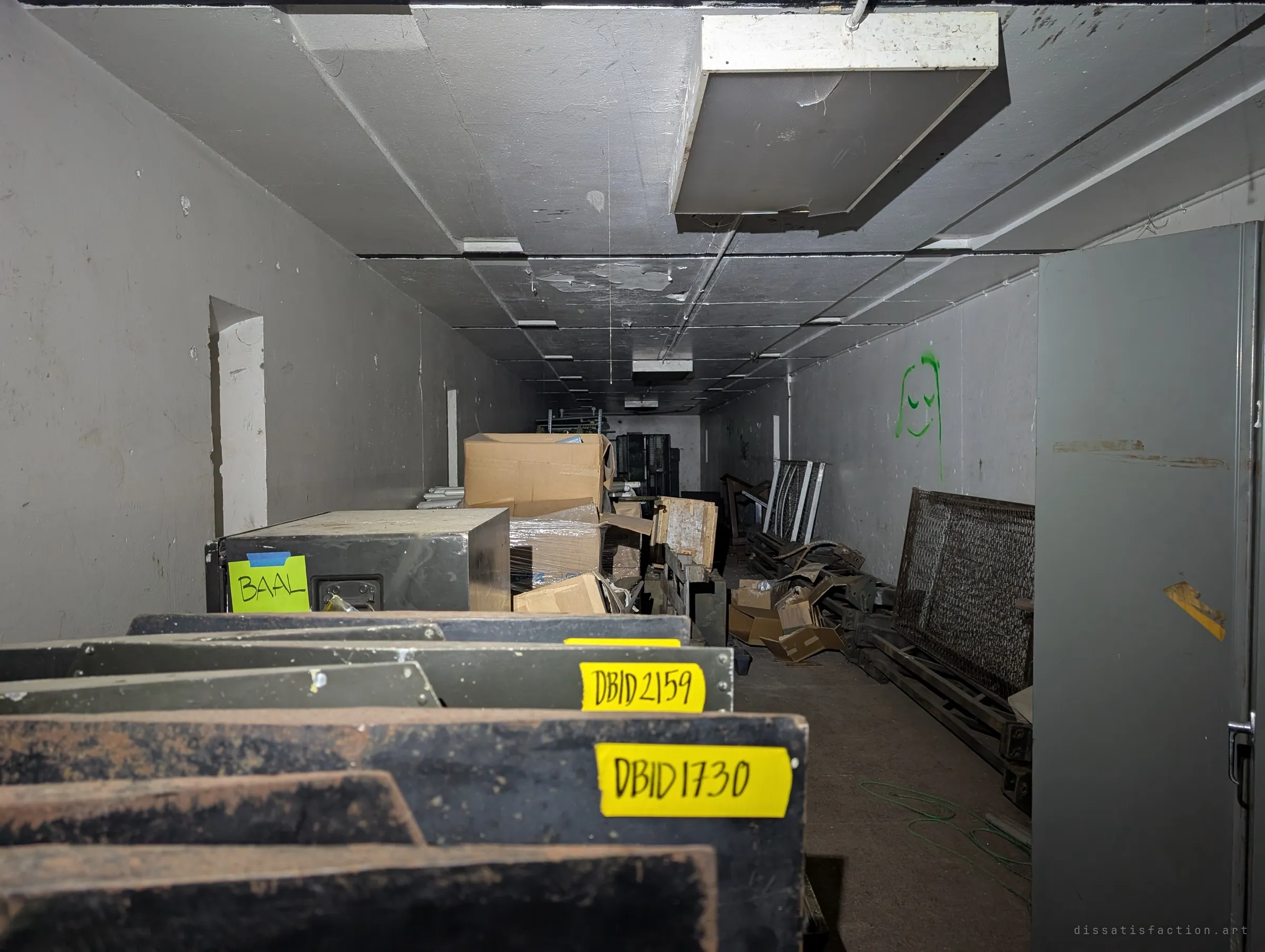 Looking over stacked olive-drab panels marked with yellow tape labels down a long storage gallery full of boxes, crates and metal grilles, a small green face drawn on the wall.