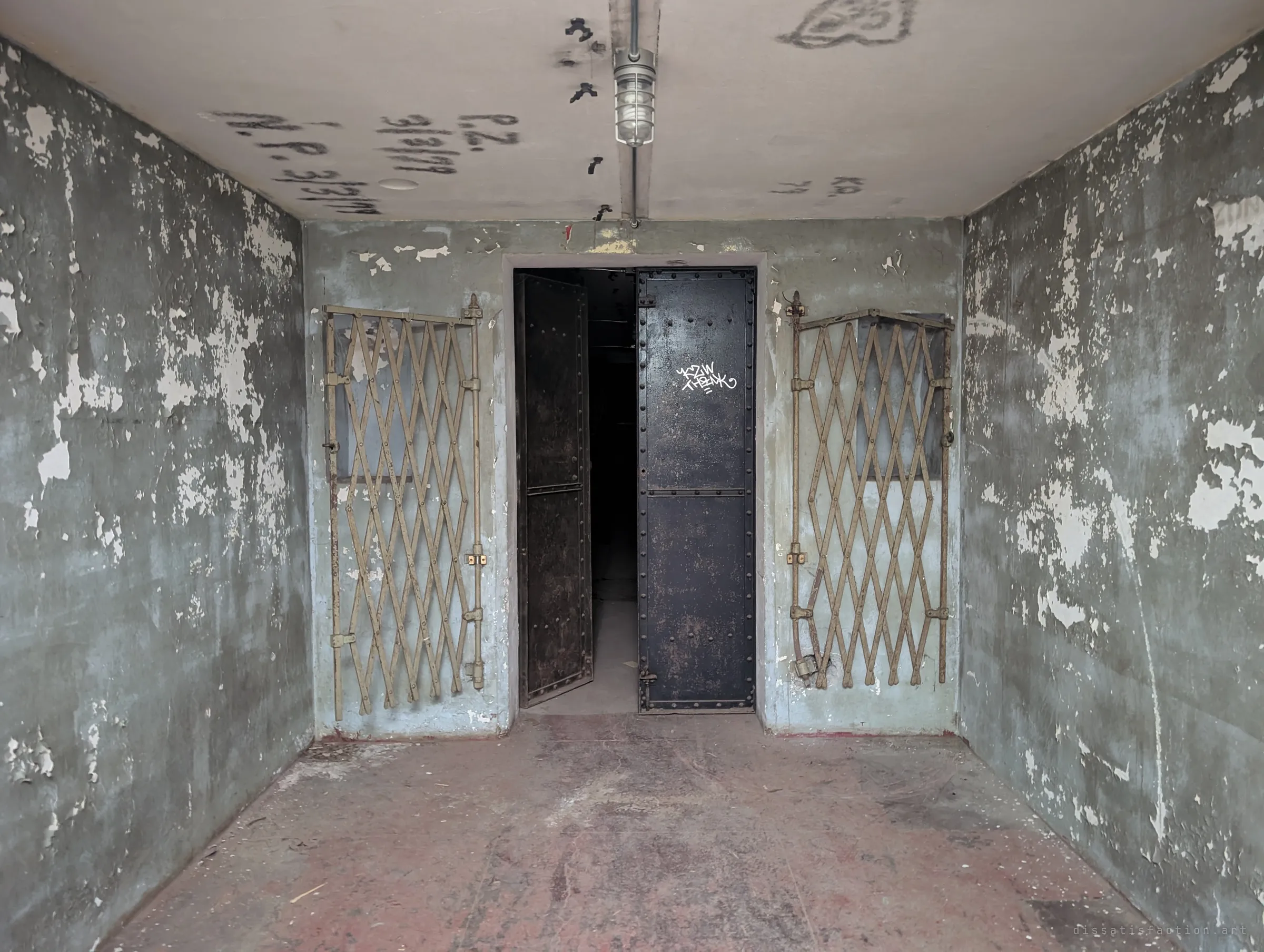 A peeling grey-green concrete chamber with smoke-written initials on the ceiling, framed by folding lattice gates on either side of a half-open riveted steel double door.