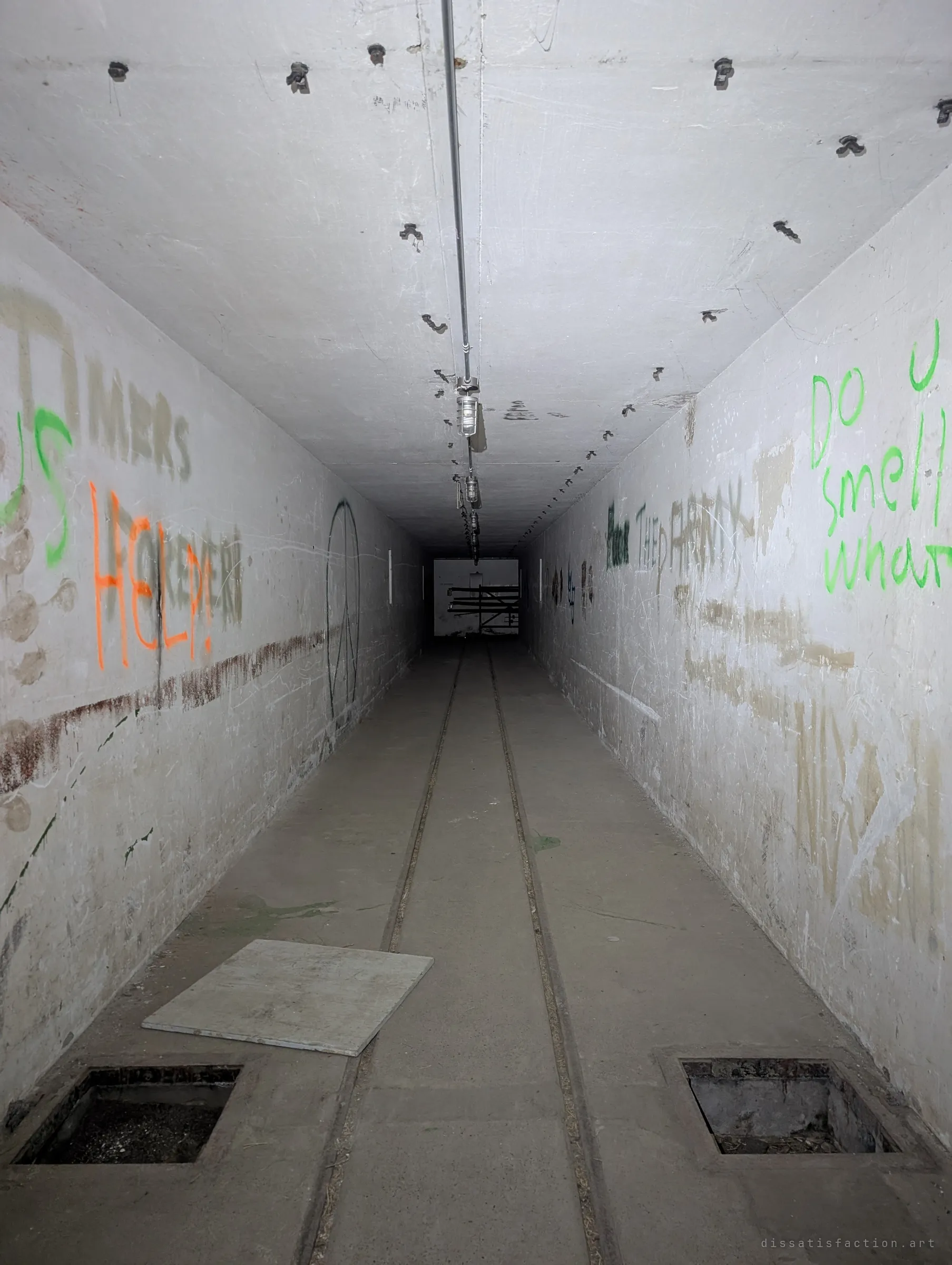 A long whitewashed concrete tunnel with rail tracks down the floor, open floor hatches and spray-painted pleas on the walls. The corridor runs straight into darkness toward a lit room at the far end.