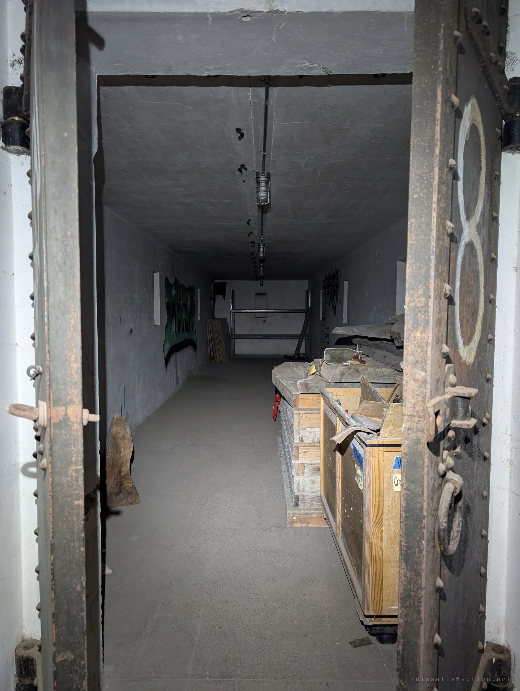 A heavy riveted steel blast door marked with a painted number stands open onto a storage gallery stacked with wooden crates and scrap timber. The door thickness shows how the space was built to take a hit.