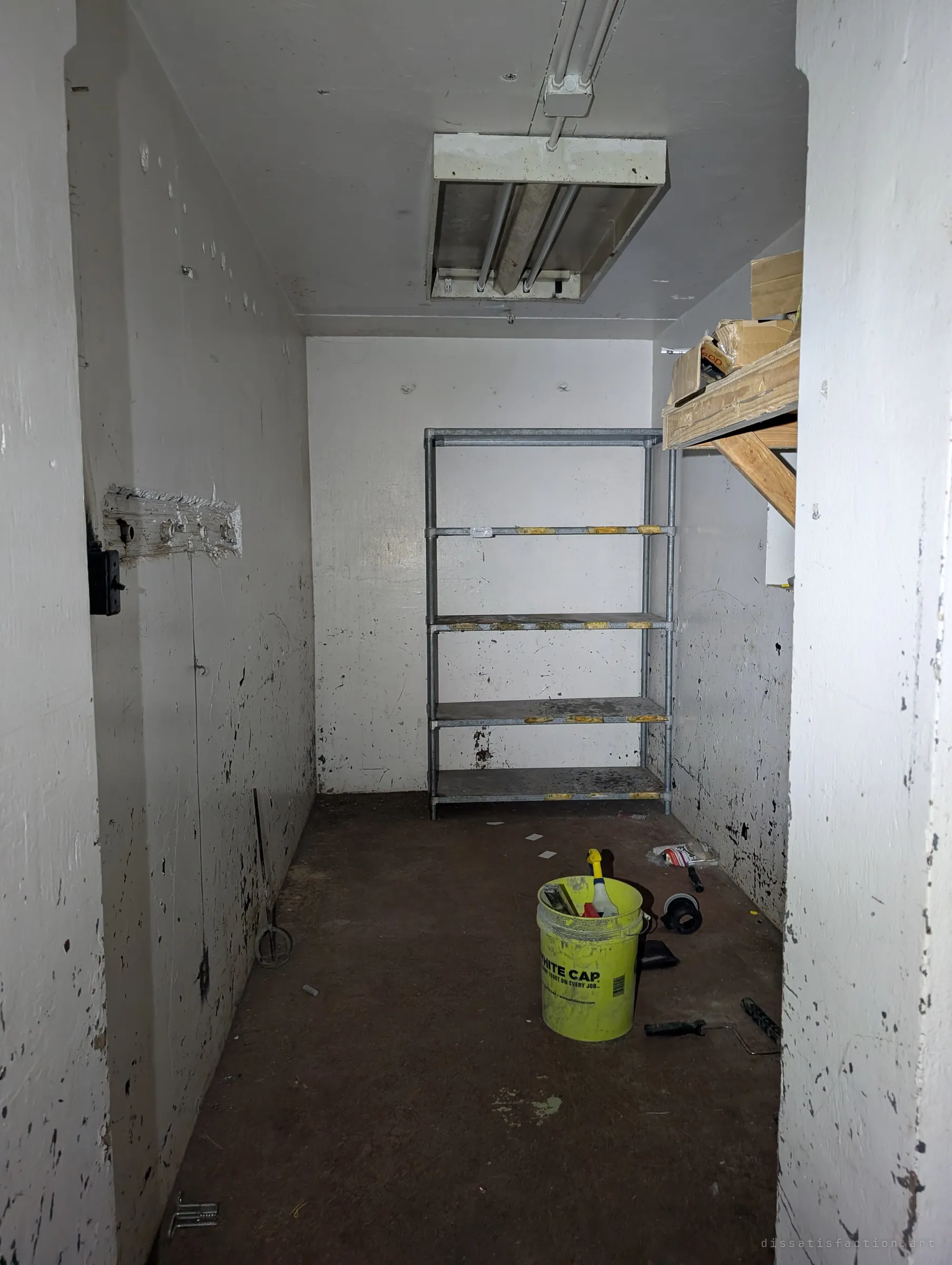 A small whitewashed storeroom with an empty wire shelf, a fluorescent fixture overhead and a yellow bucket of tools left on the floor. It reads as a space someone was still maintaining not long ago.