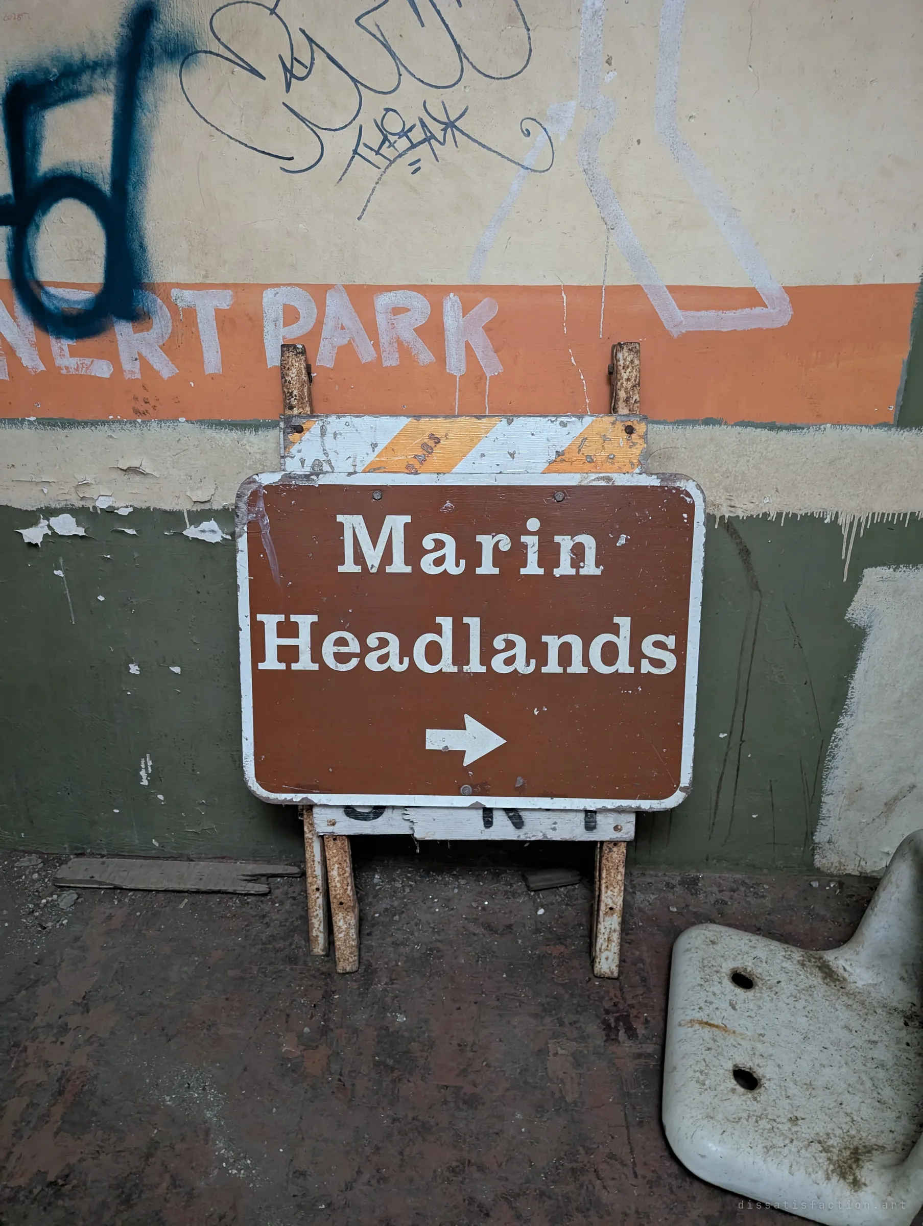 A weathered brown directional sign propped on an old barricade against a graffiti-marked wall, beside a broken porcelain fixture. A leftover from when the place still had visitors to direct.