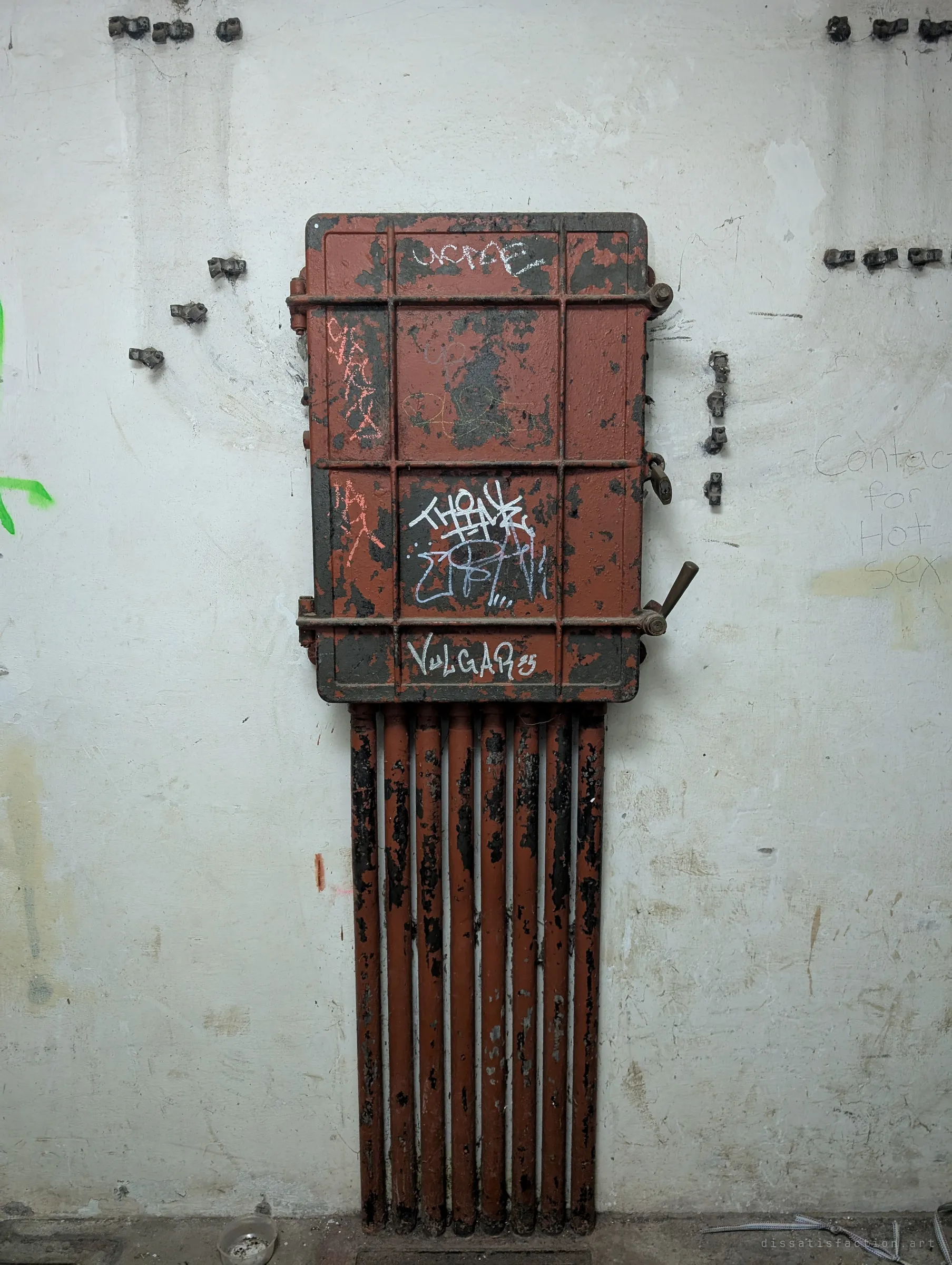 A rusted cast-iron hatch with a bank of pipes running beneath it, tagged with graffiti on a white bunker wall. The old ventilation or heating hardware still hangs where it was bolted in.