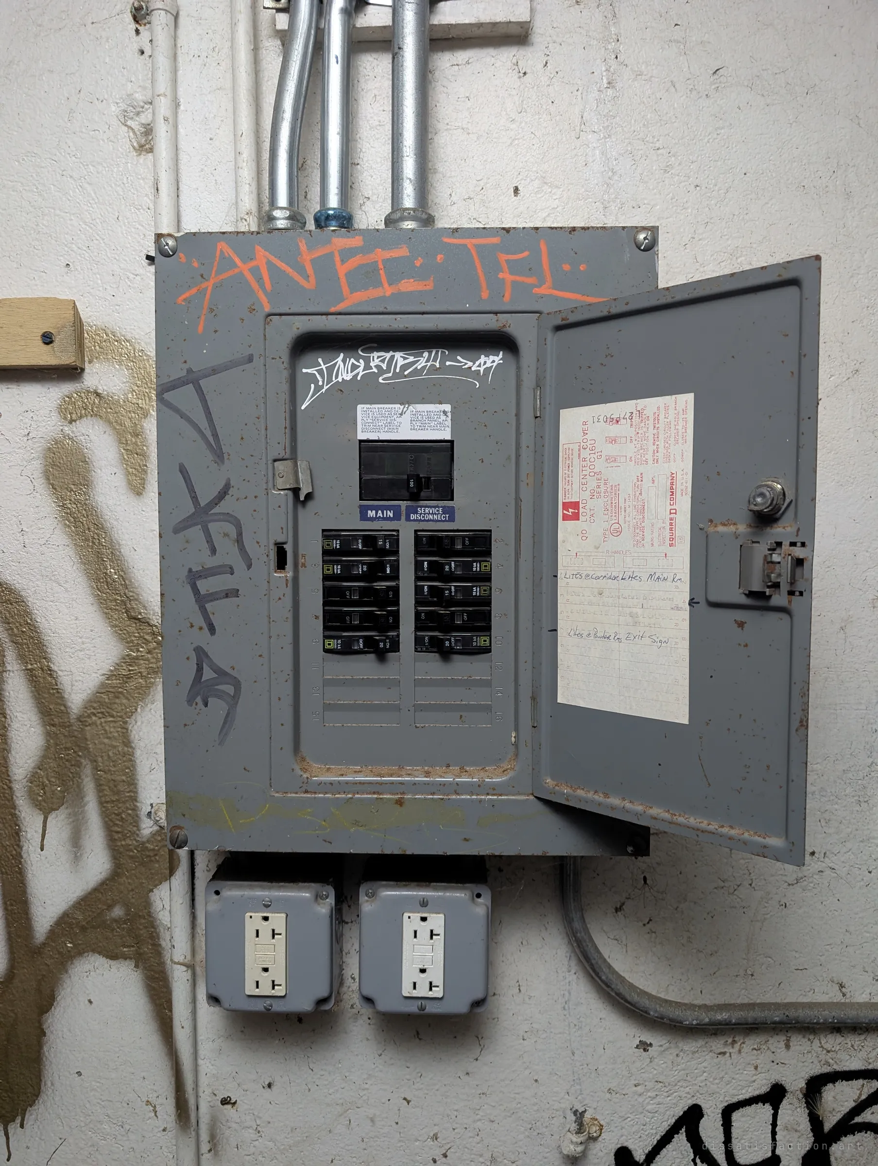 An open grey breaker panel with a handwritten circuit card for corridor and room lights, two outlets below and graffiti around it. Later wiring added to a much older concrete structure.