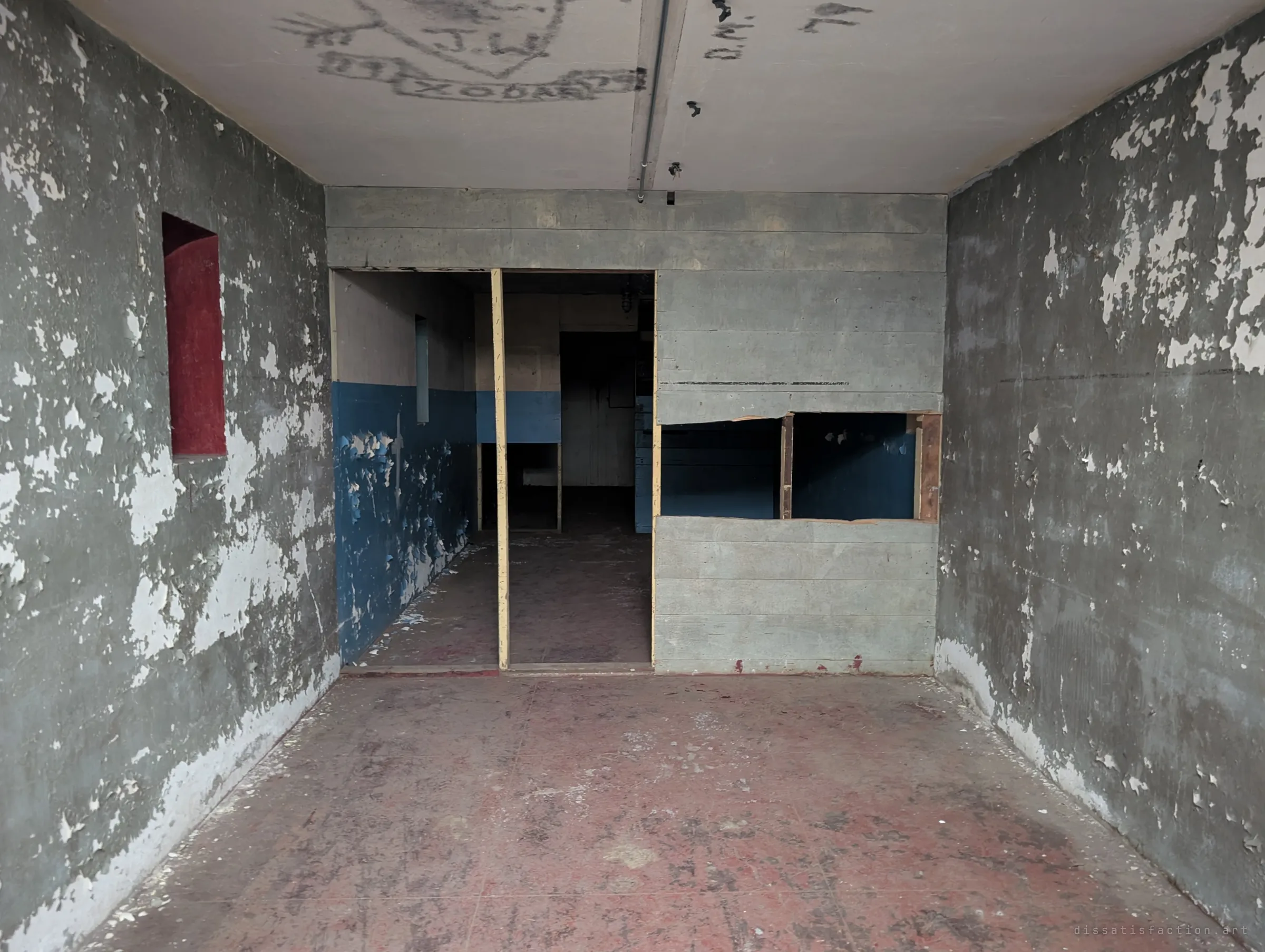 A concrete room with peeling grey and blue paint, a red recessed niche and a wooden partition opening onto further rooms. Layers of paint flake away to show decades of reuse.