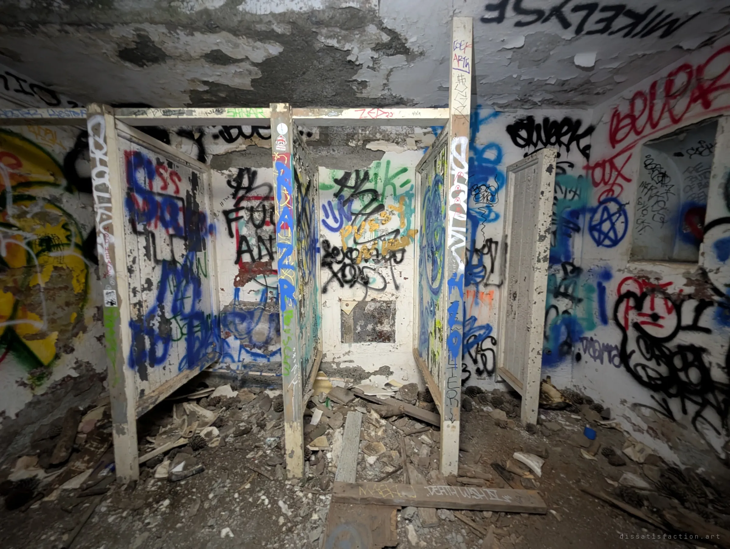A row of wooden stall partitions in a rubble-strewn room, every surface tagged with spray paint. The old latrine layout is still clear under the graffiti.