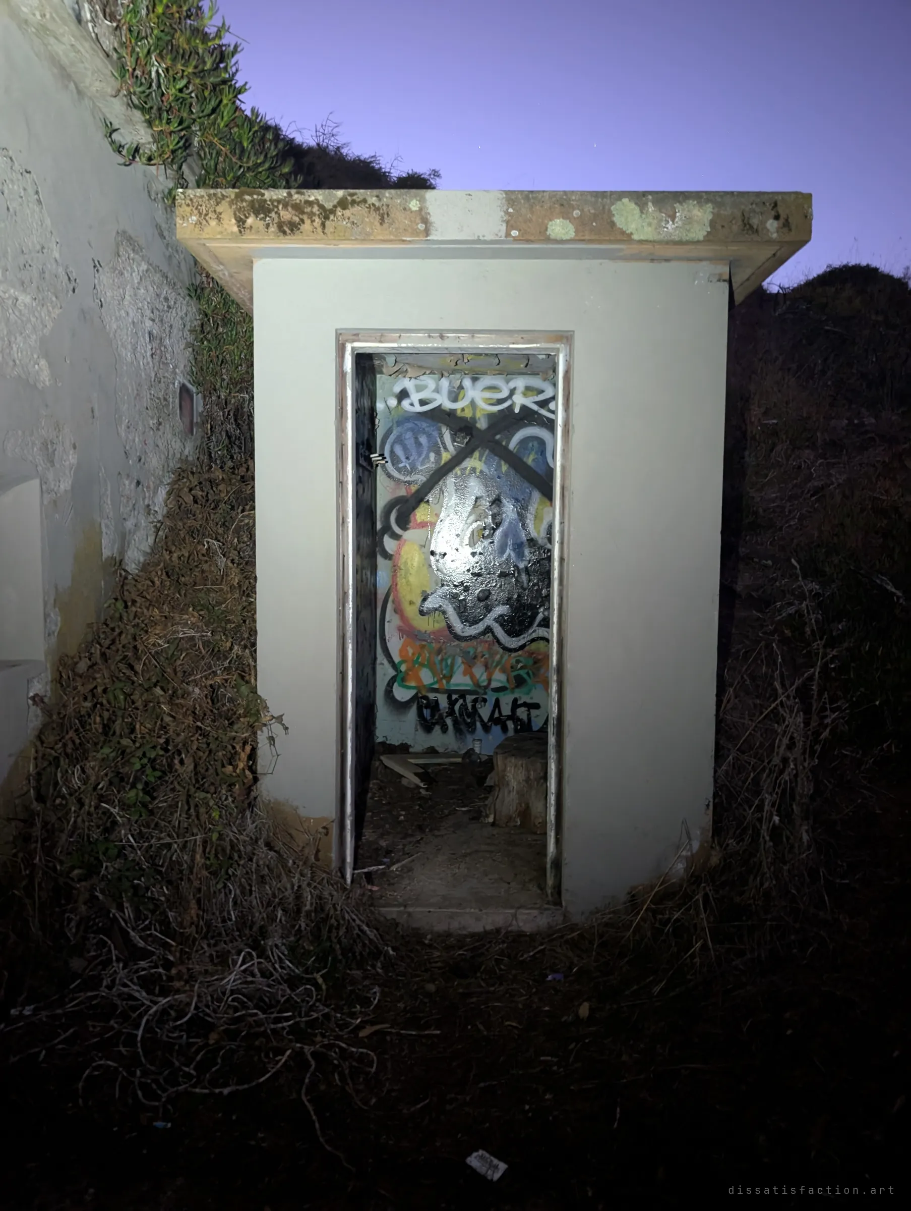 A small concrete entrance hut set into an overgrown earth berm at dusk, its doorway lit to show graffiti inside. The outside view shows how the structure is buried into the hill.