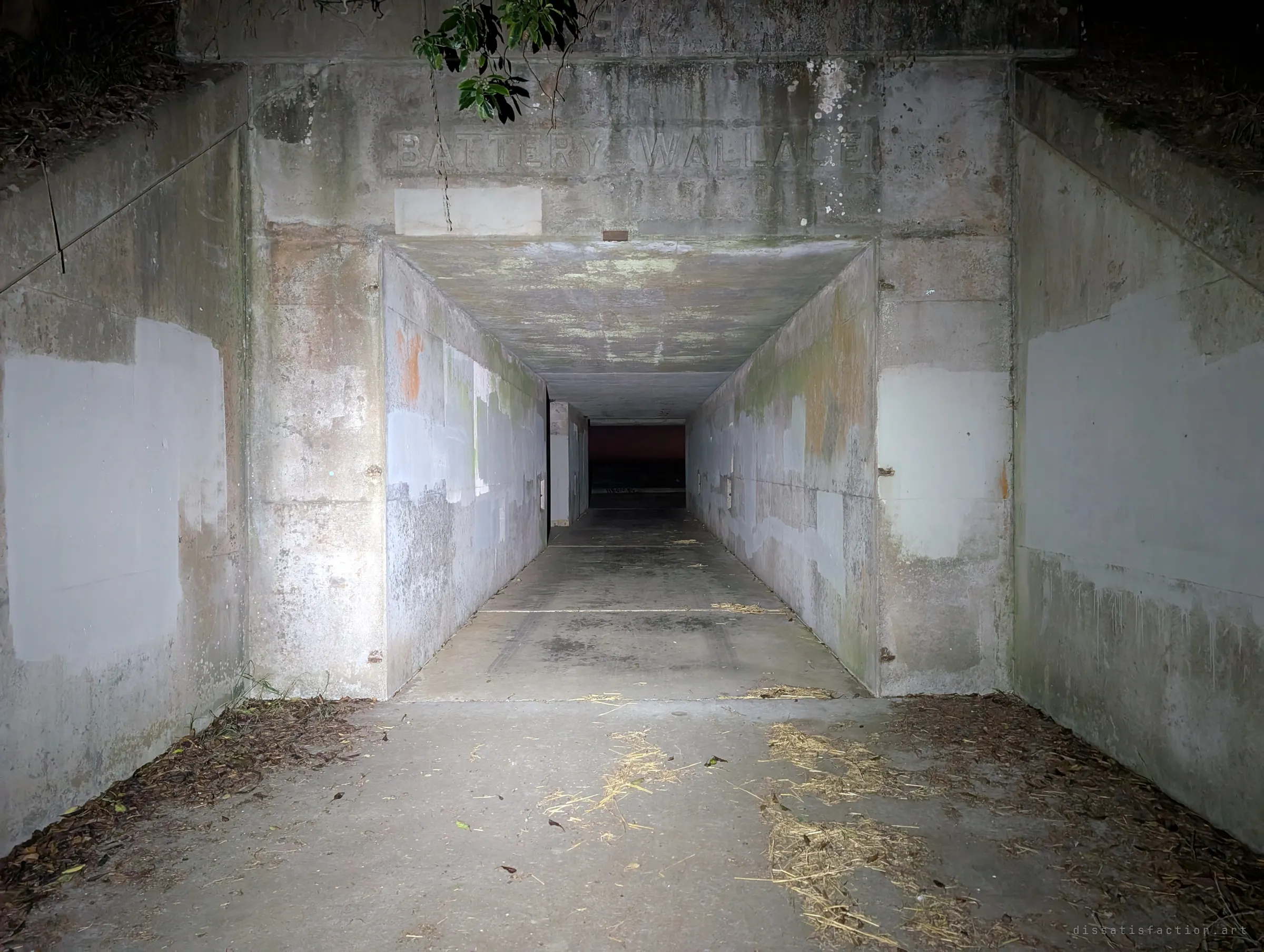 A long, flashlit concrete passage runs straight into a coastal gun battery, its walls patched in mismatched grey paint. The lettering above the portal has been blurred.
