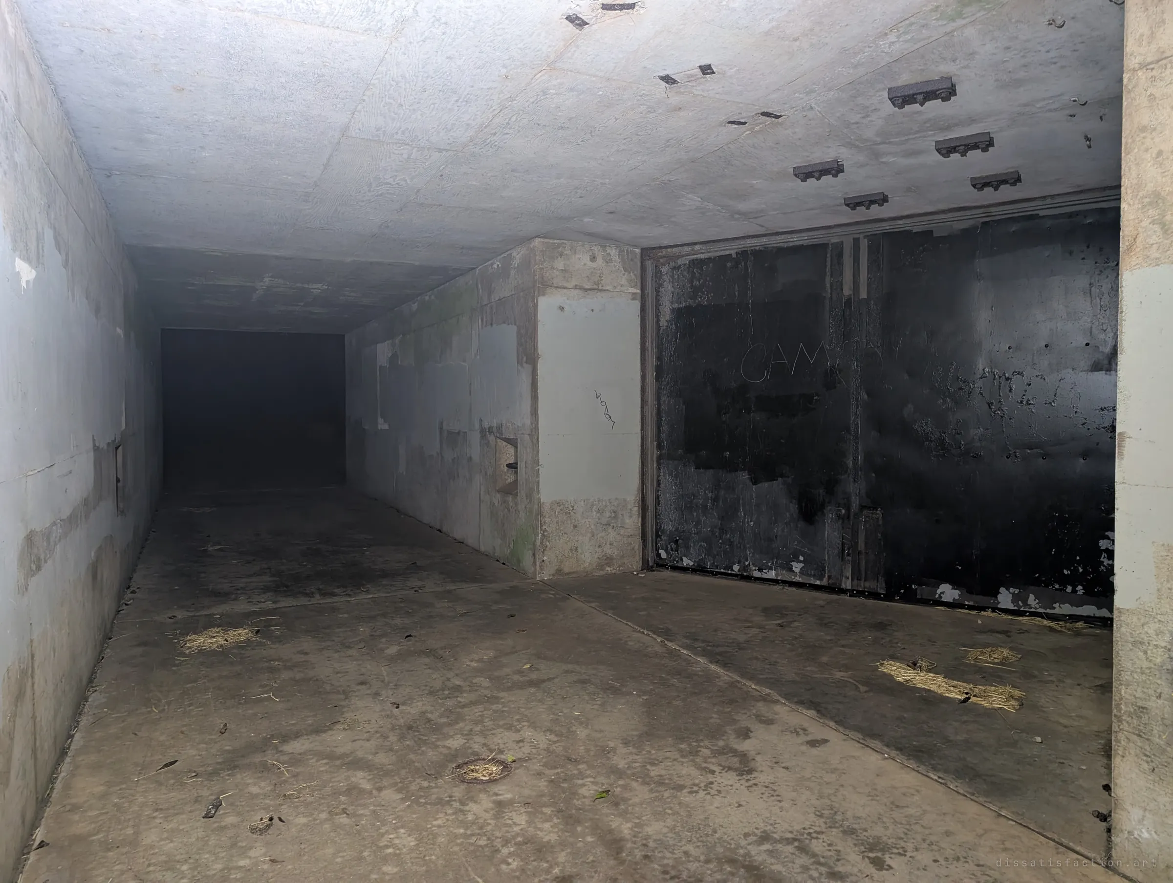 A wide corridor inside the battery meets a blackened steel blast door scratched with graffiti. Rusted ceiling brackets and scattered straw mark decades of disuse.