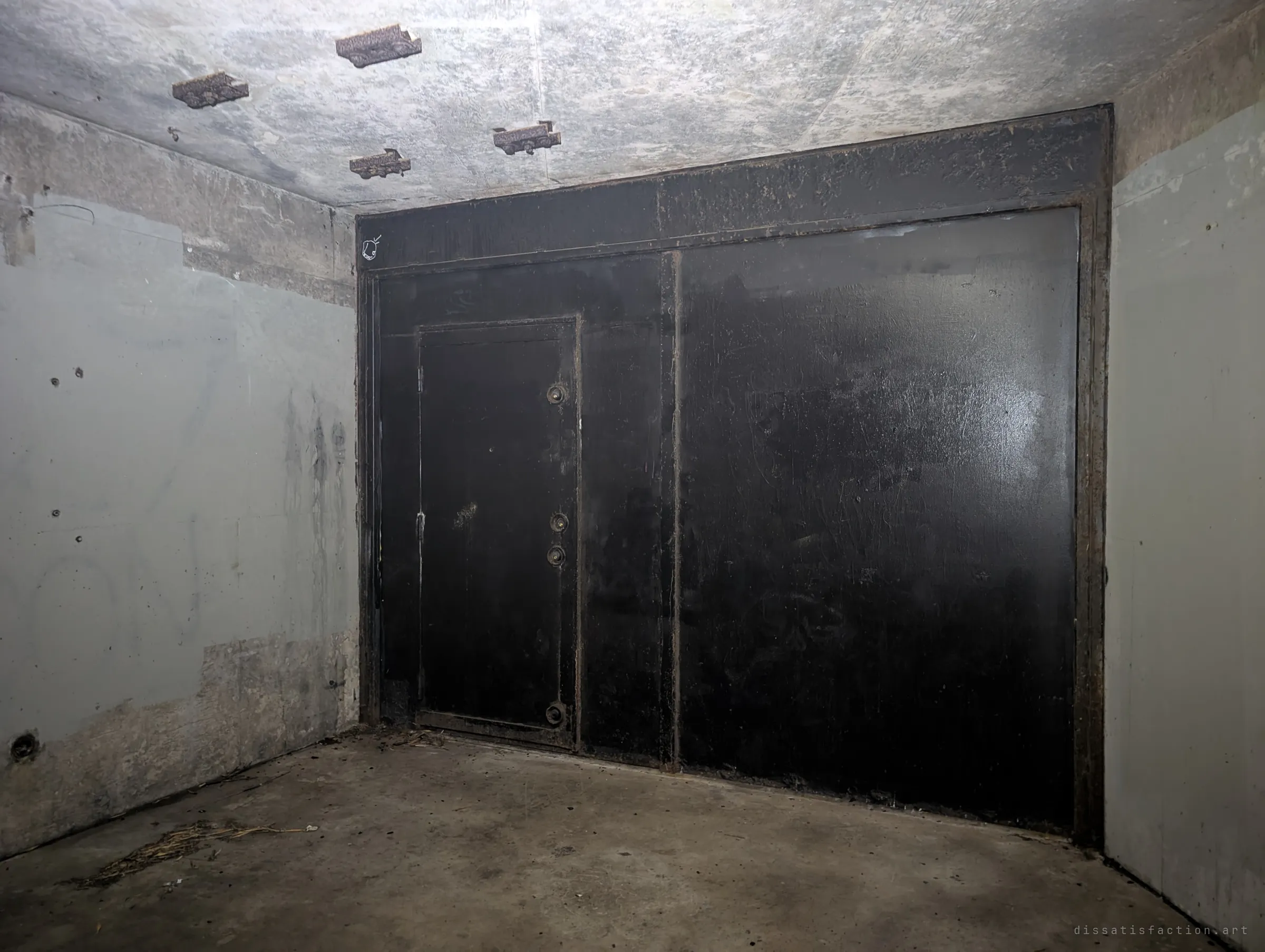 A heavy black steel wall with a riveted inset door seals off a magazine room, its grey walls peeling around it. Rusted brackets still hang from the ceiling.