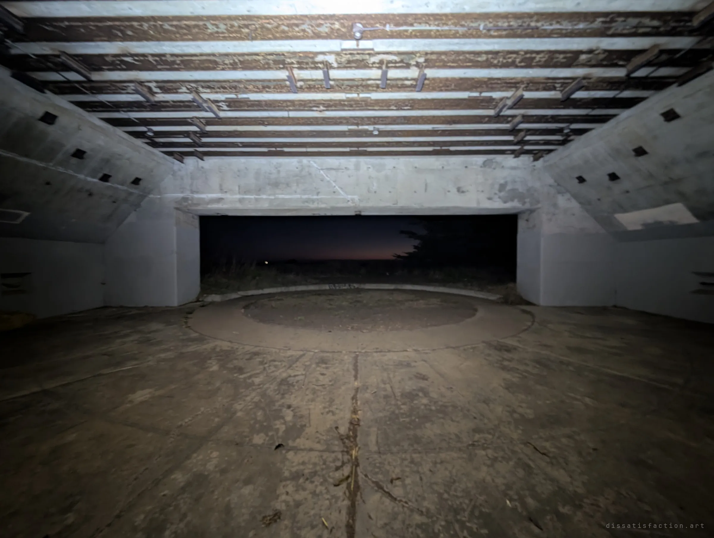 An ultra-wide view under the steel-ribbed roof of a gun emplacement, with the circular mount in the floor and a last band of dusk glowing through the opening. It shows the scale of the casemate that once held the gun.