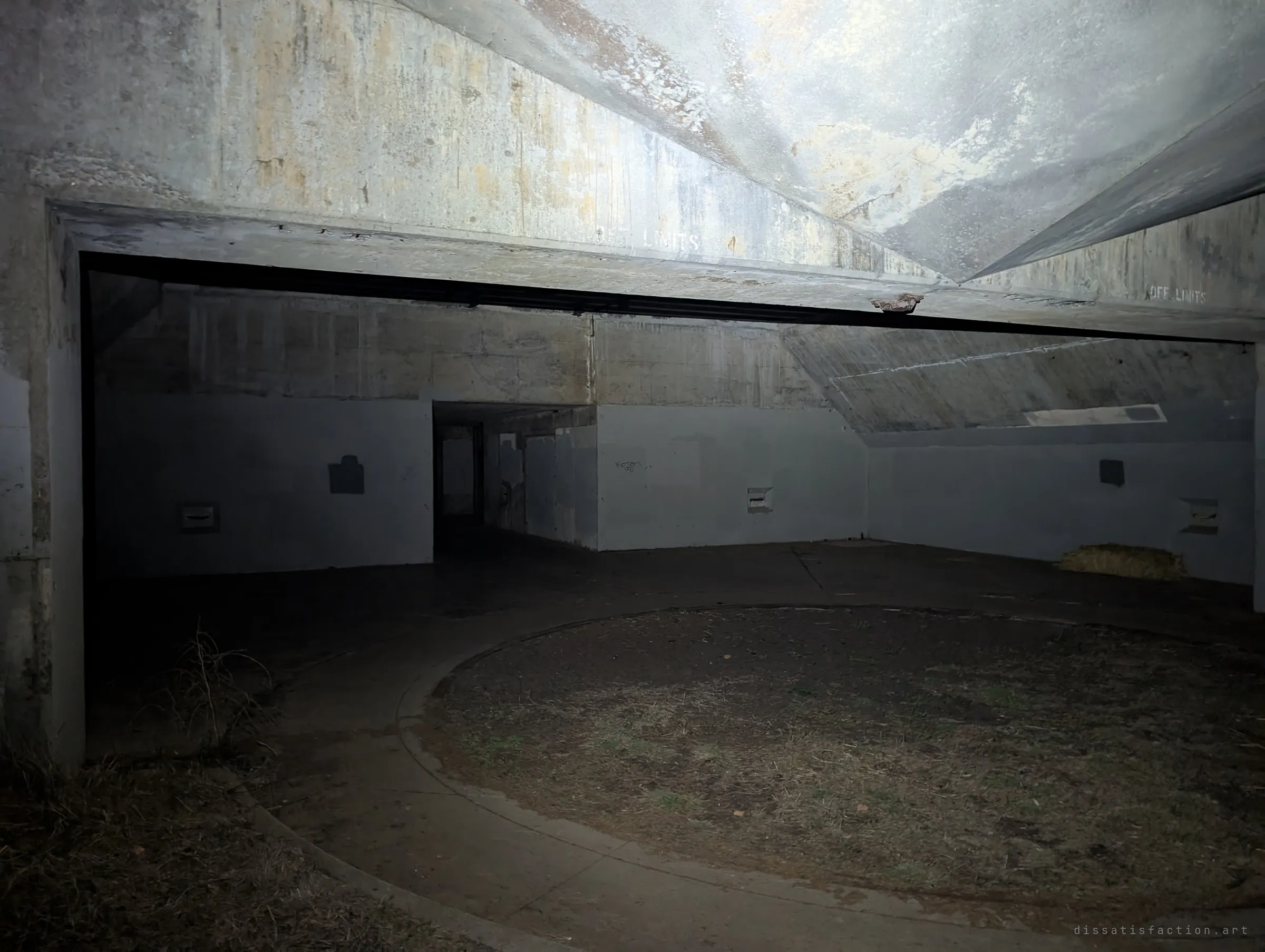 The circular gun pit sits in shadow beneath a massive angled concrete canopy, with a dark doorway leading deeper into the battery. Faded stencilled warnings line the beam overhead.