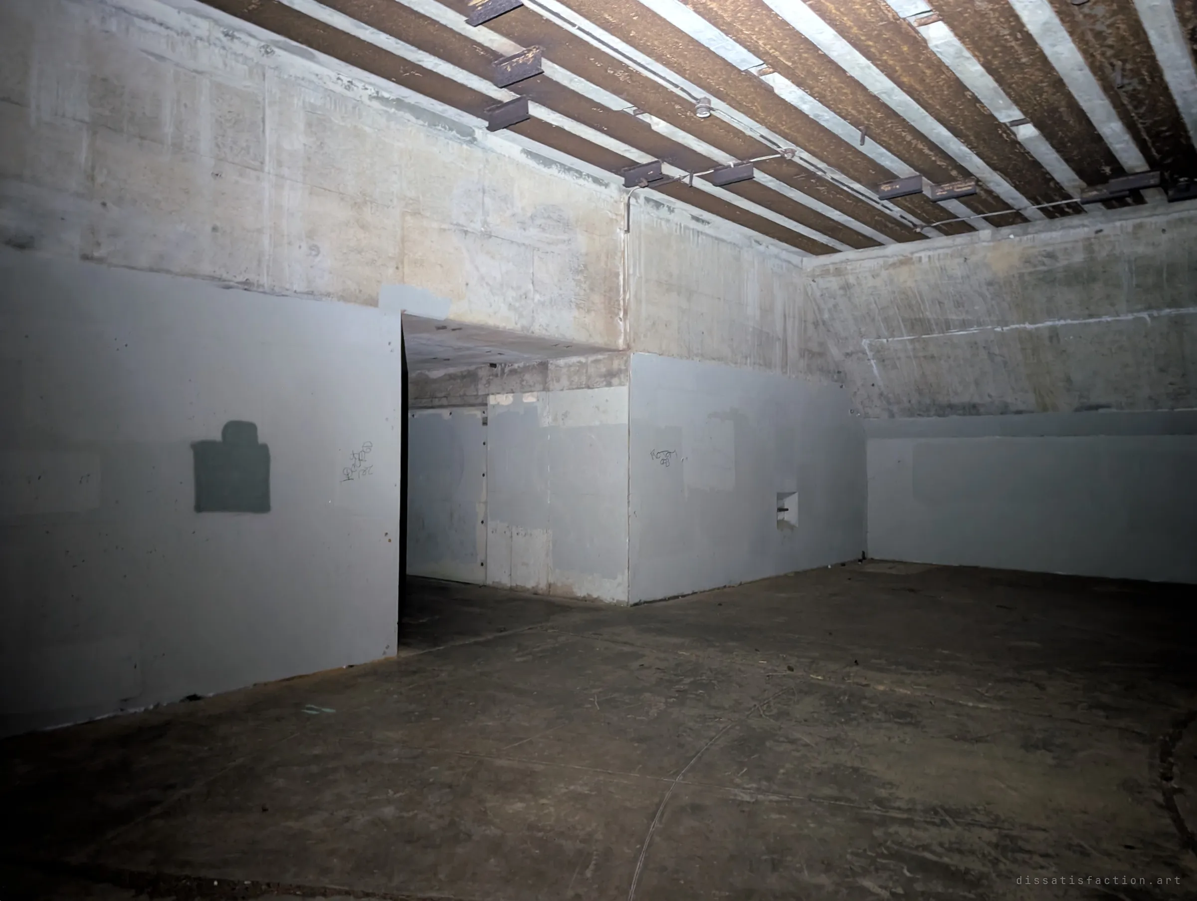 Inside the emplacement, rusted steel ribs stripe the ceiling above grey painted walls and a passage leading back into the hill. Small tags and a painted-over patch are the only marks of visitors.