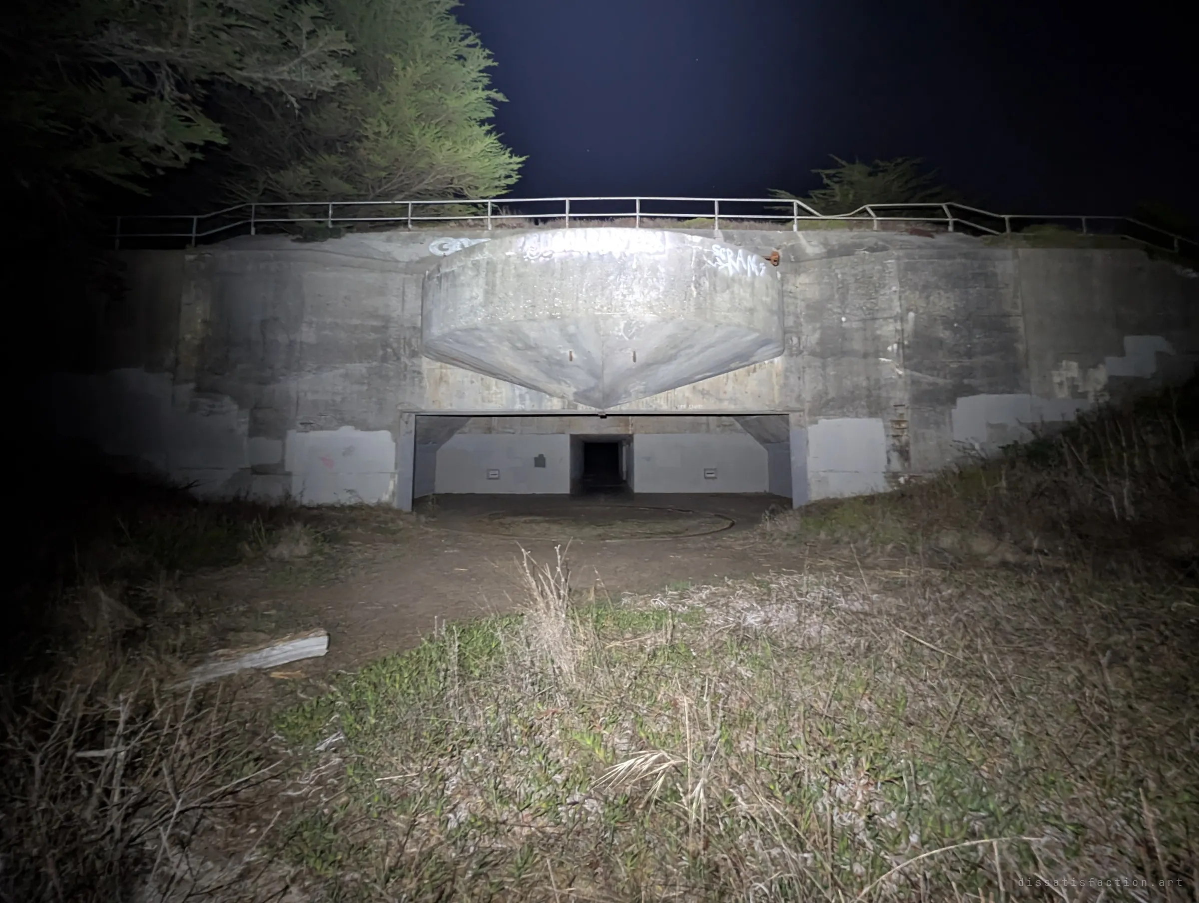 The battery seen from outside at night: a heavy concrete shield juts over the wide gun opening, tagged with graffiti, with a railing and cypress trees silhouetted above. It shows how the whole emplacement is buried into the hillside.