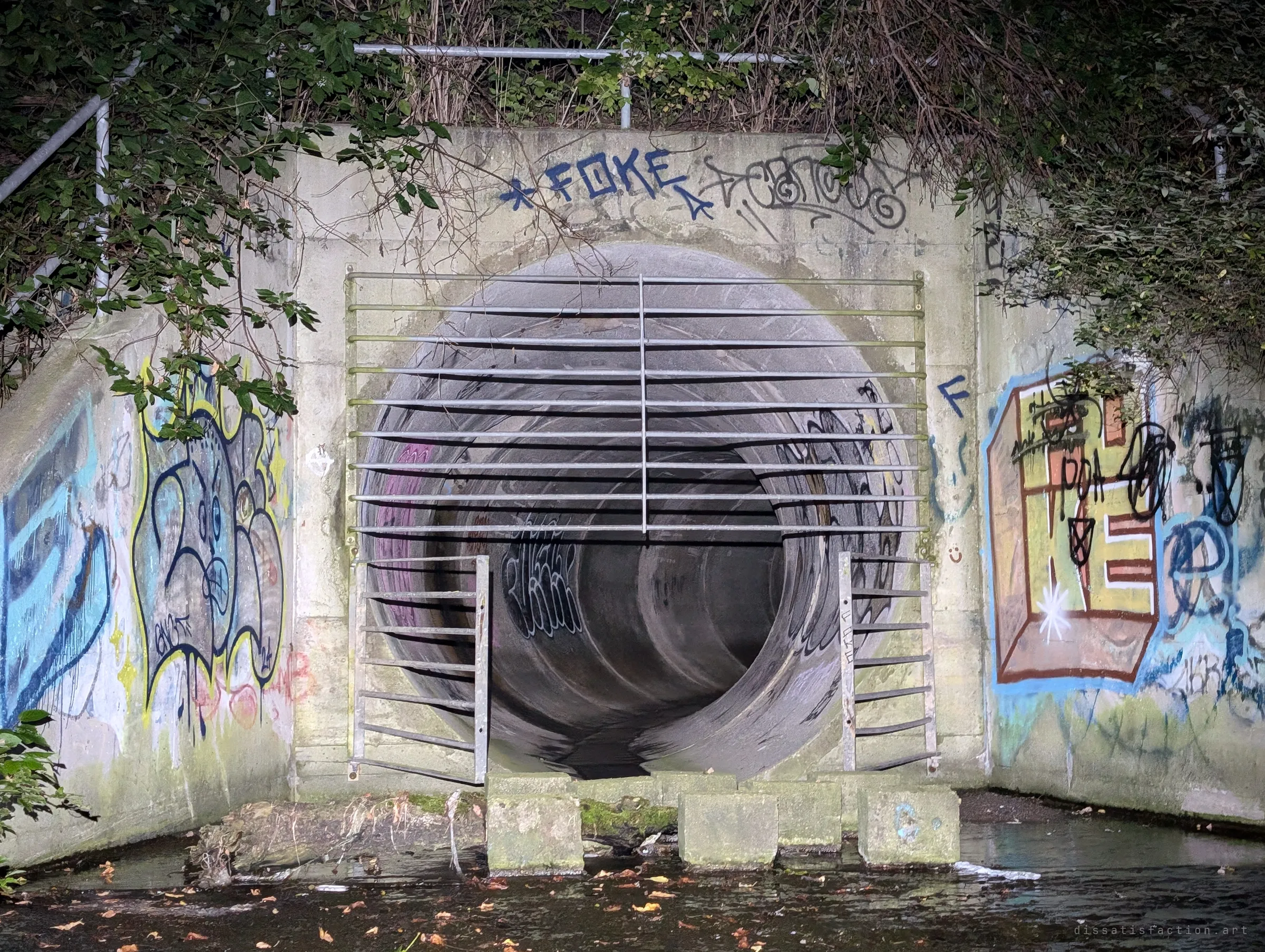Head-on view of the barred outfall, horizontal bars across a large concrete pipe flanked by colourful graffiti and stepping blocks in the water. The gate is the way in.