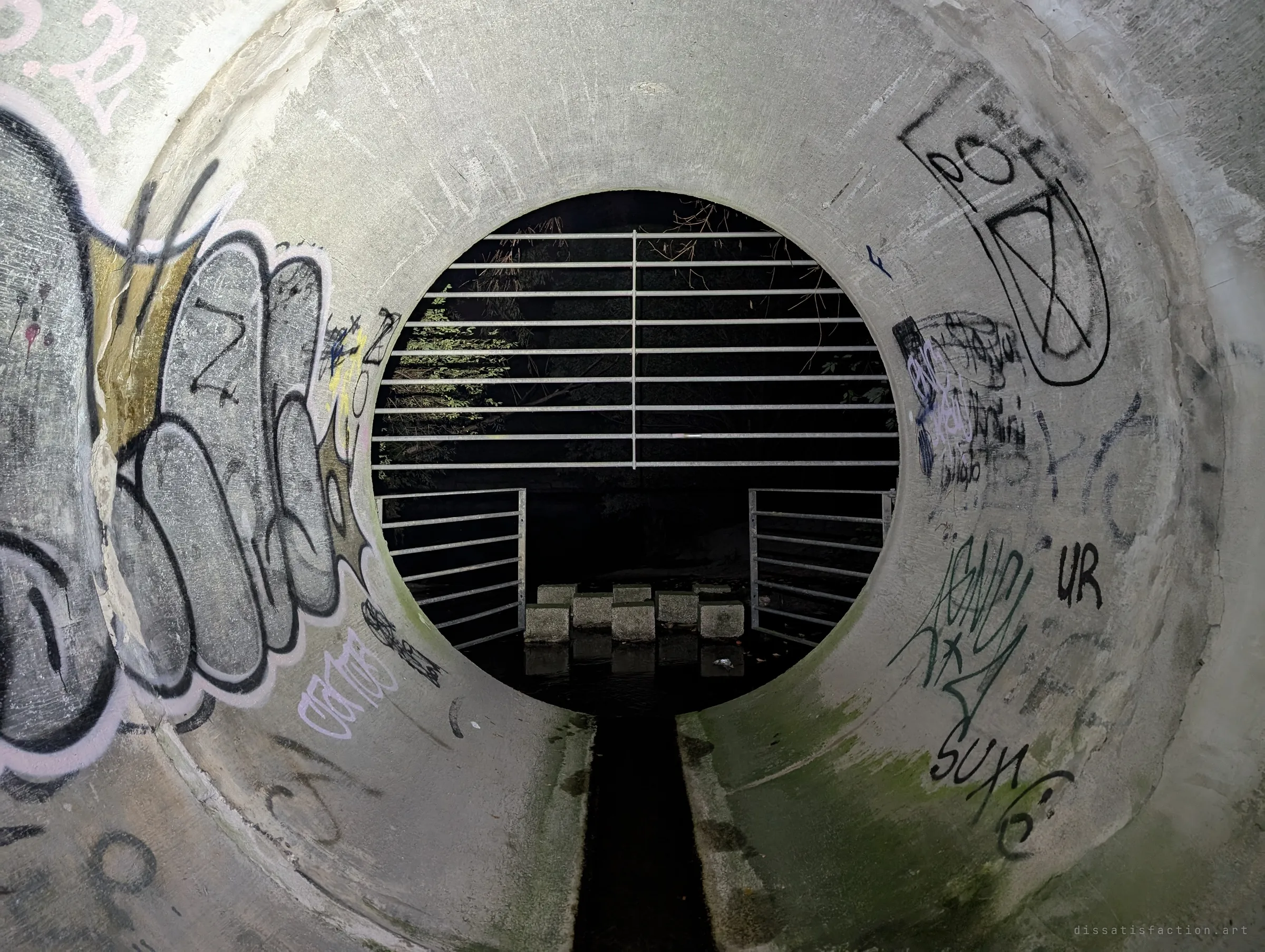 Looking back out from inside the pipe toward the barred entrance, the tunnel walls thick with tags and a narrow dark channel running down the middle.
