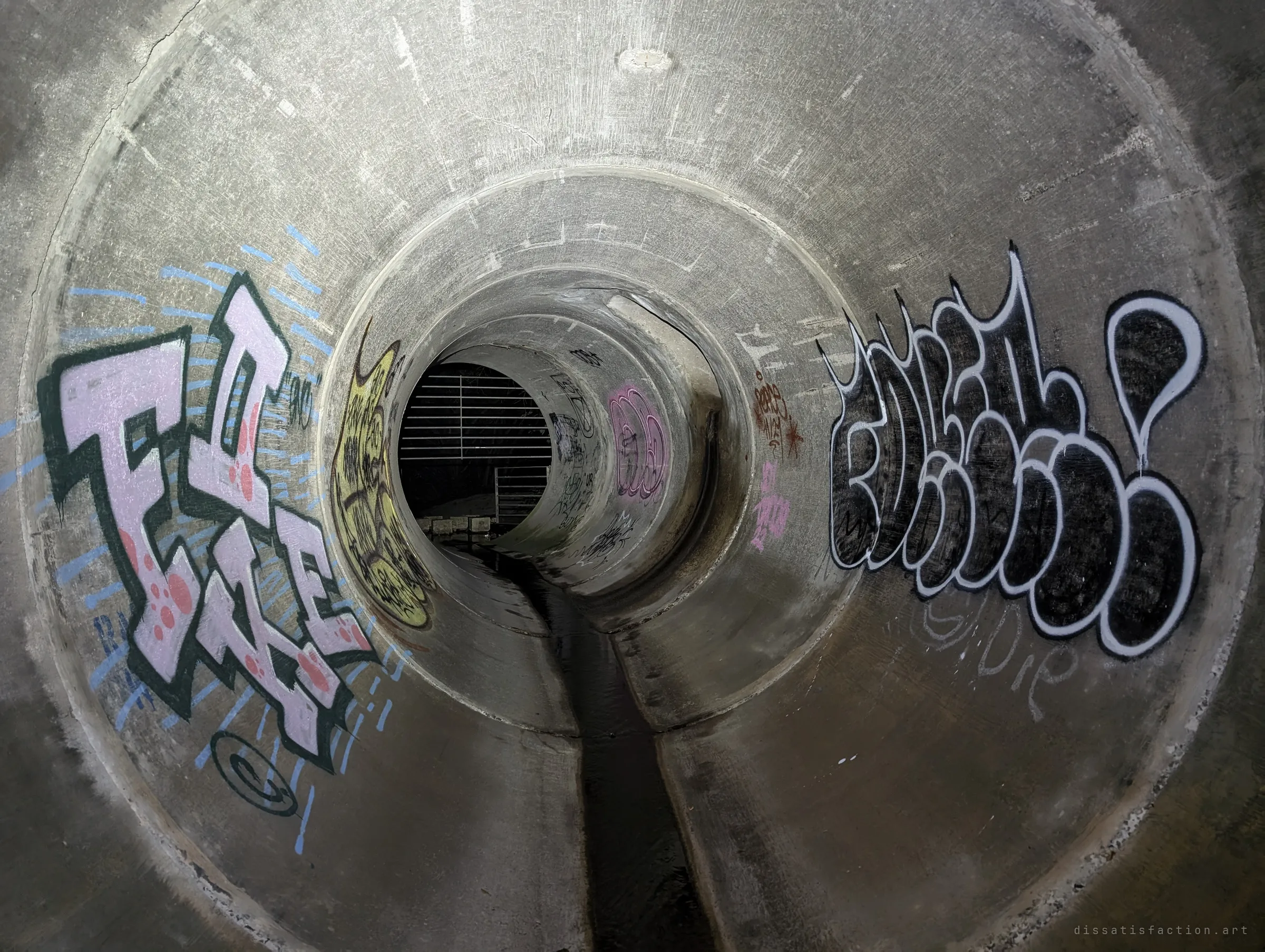 Large graffiti pieces on both walls of the concrete pipe, which telescopes back to the distant grated entrance. A thin trickle runs along the low-flow channel.