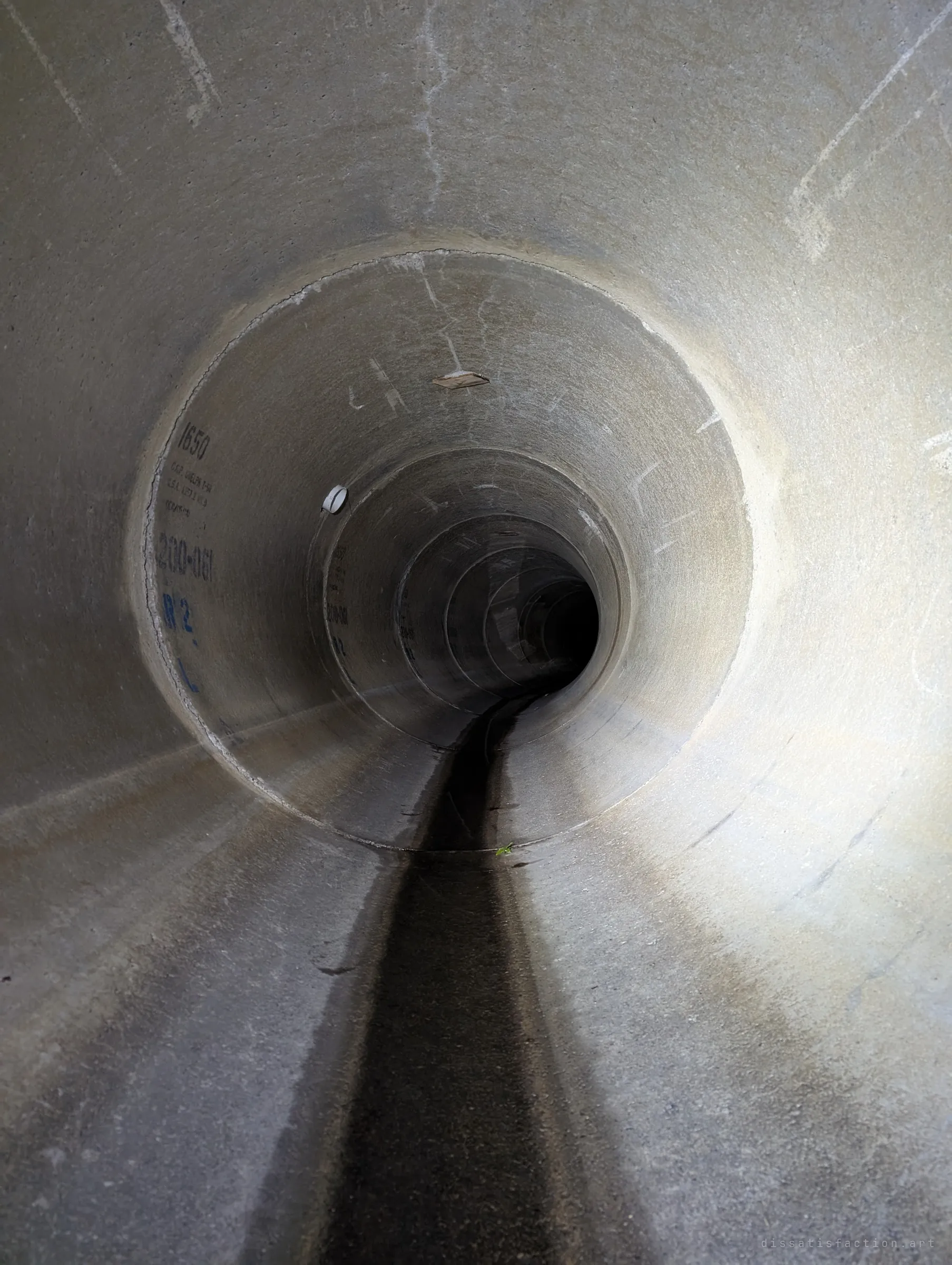 A clean, bare concrete pipe stretching into darkness, segment joints ringing away toward a black vanishing point with a wet channel along the floor. Only stencilled pipe specs mark the walls.