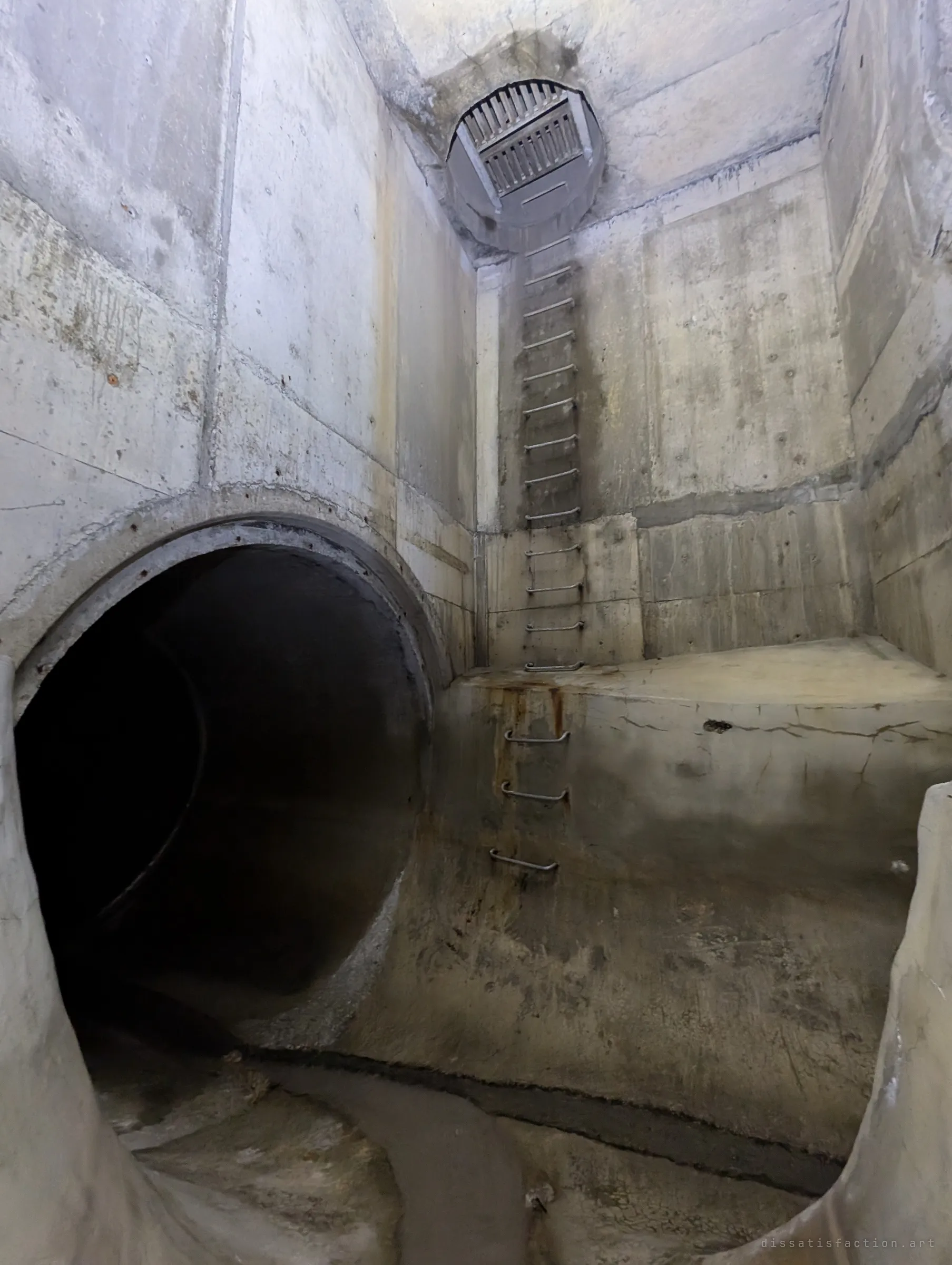 A tall concrete junction chamber with a pipe opening at its base and a ladder of rungs climbing to a grated shaft far above. The first glimpse of daylight access from below.