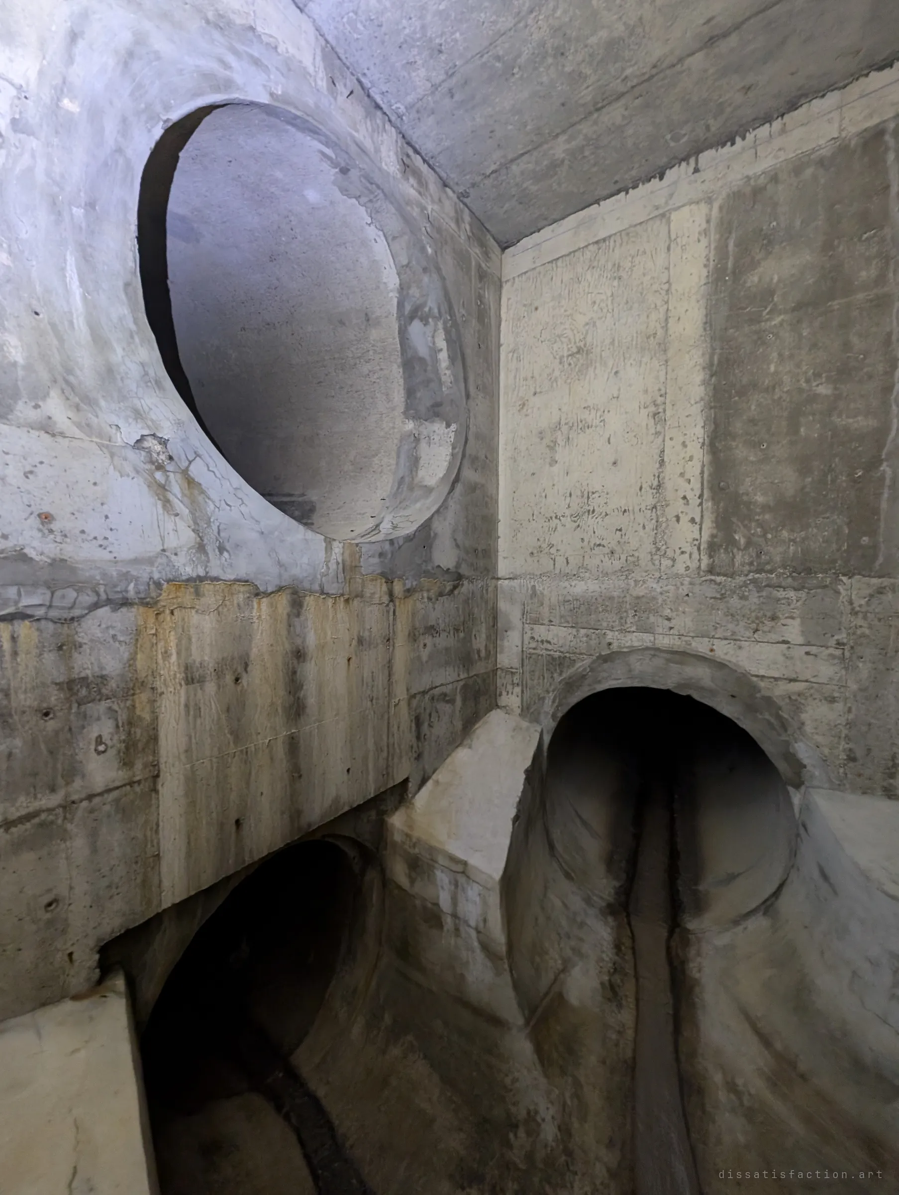 Three concrete pipes meet in a stained chamber: one large bore high on the wall and two lower inlets, each with a channel carved into the floor. The left pipe carries an illegal sanitary connection from upstream.