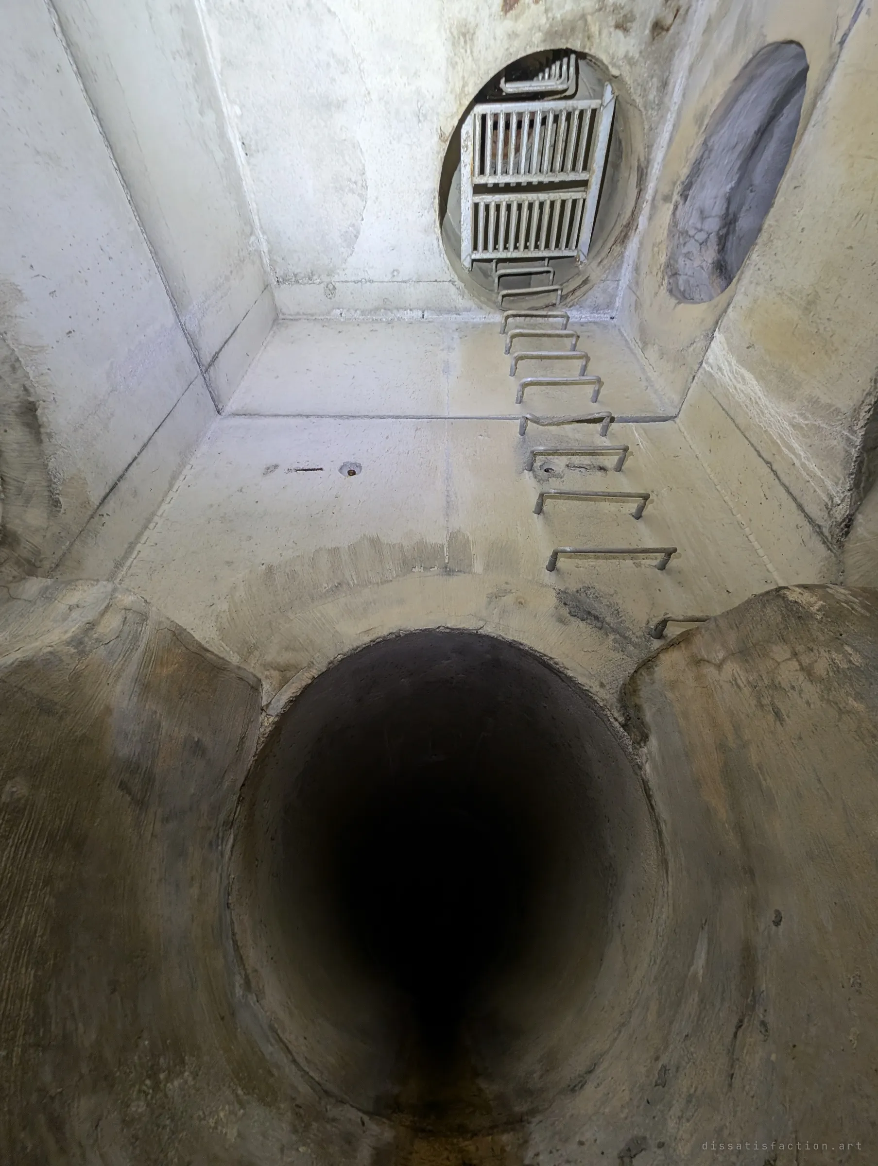 Looking straight up a concrete shaft past a pipe mouth, rungs climbing the wall to a pair of steel grates at the top. The way out, seen from the bottom.