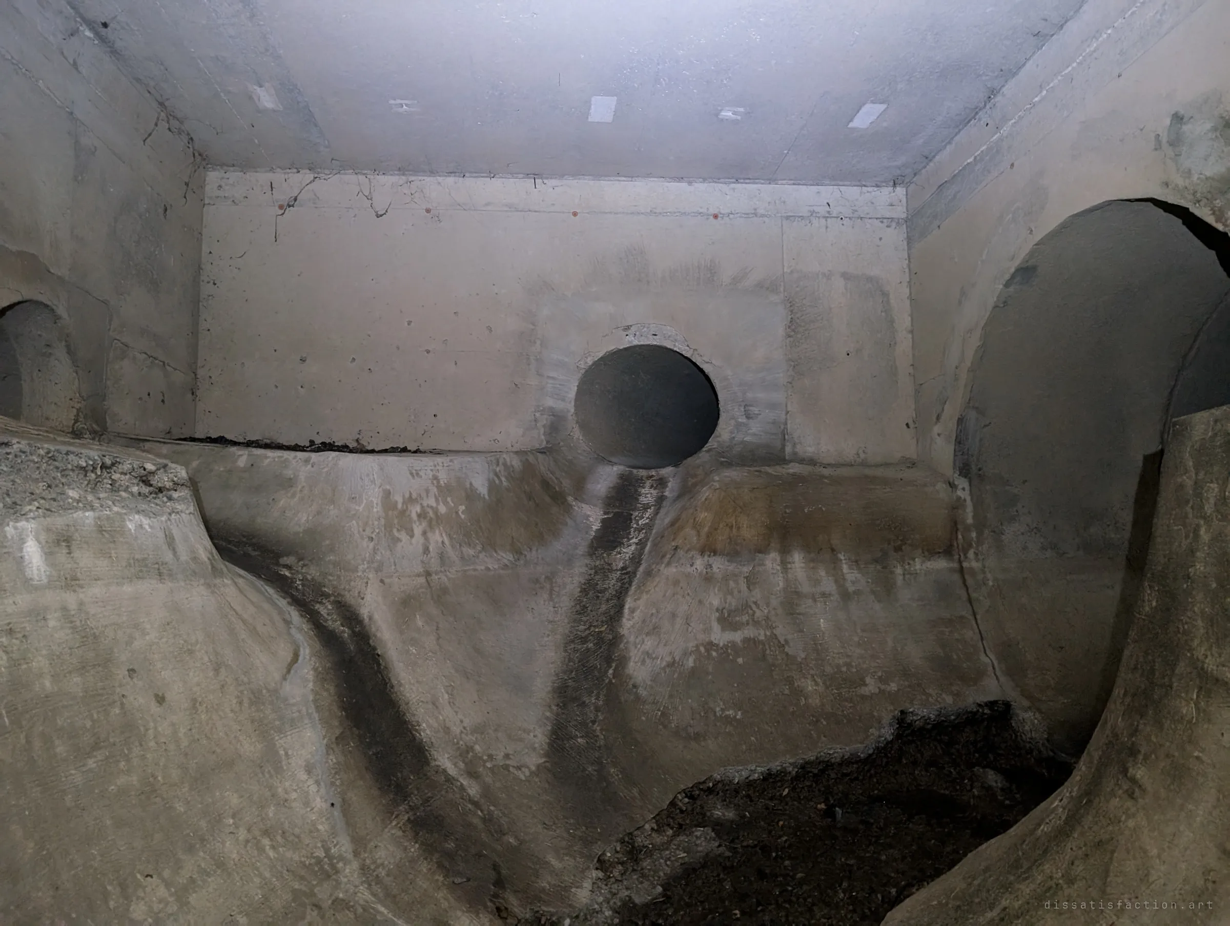 A concrete junction chamber where a small round pipe spills a dark water stain down a sculpted benching between two larger tunnel mouths. The smooth, carved floor shows how several flows are merged underground.