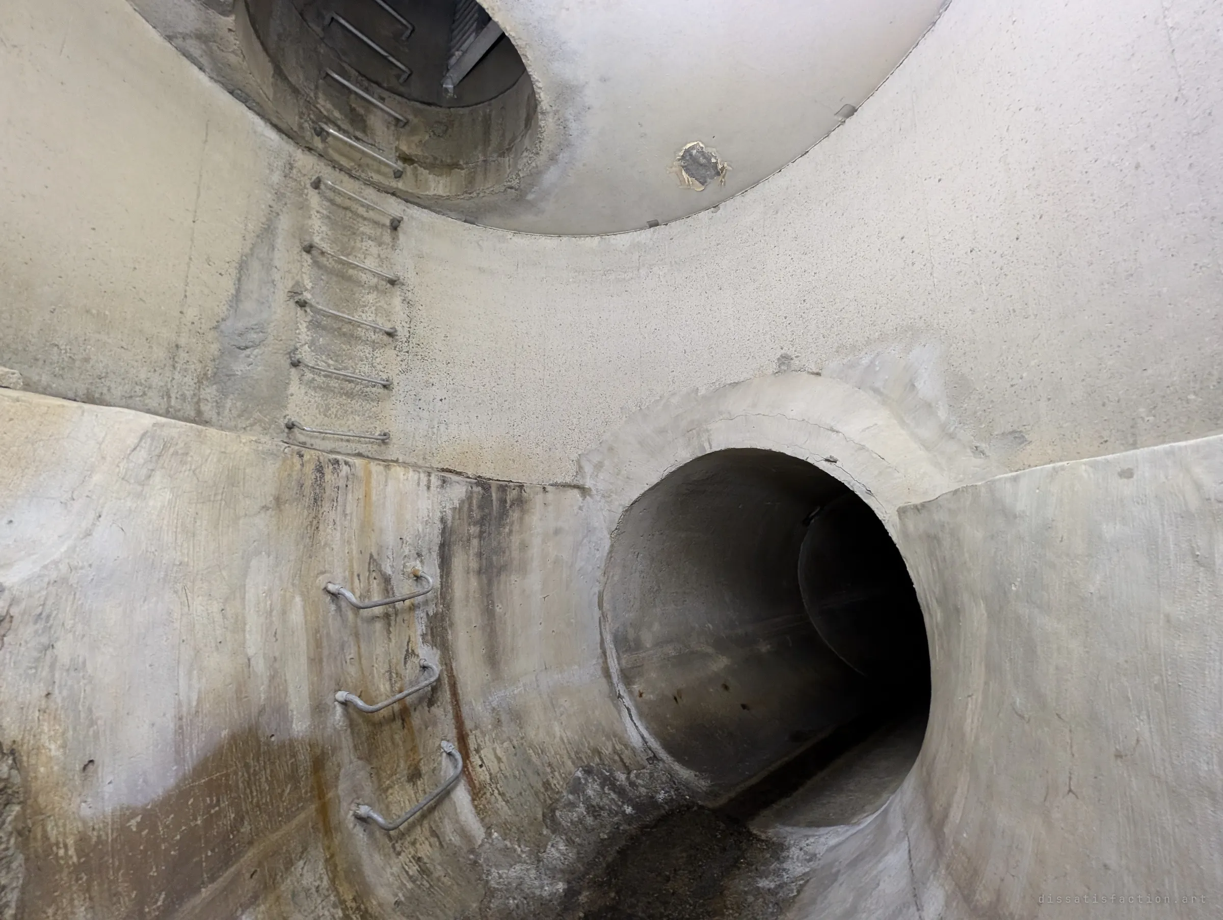 Looking up inside a round concrete shaft: steel ladder rungs climb the wall toward an access opening, while a large pipe mouth leads off into the dark below. The curving walls show the scale of the junction.