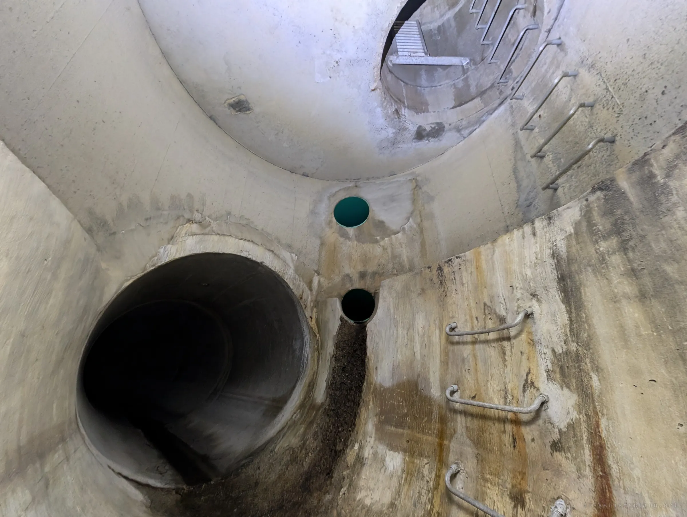 A wide view of the same shaft from another angle, with a big pipe opening on the left, two small green-lined inlets in the middle and rust-streaked ladder rungs on the right. Light falls in from the access opening above.